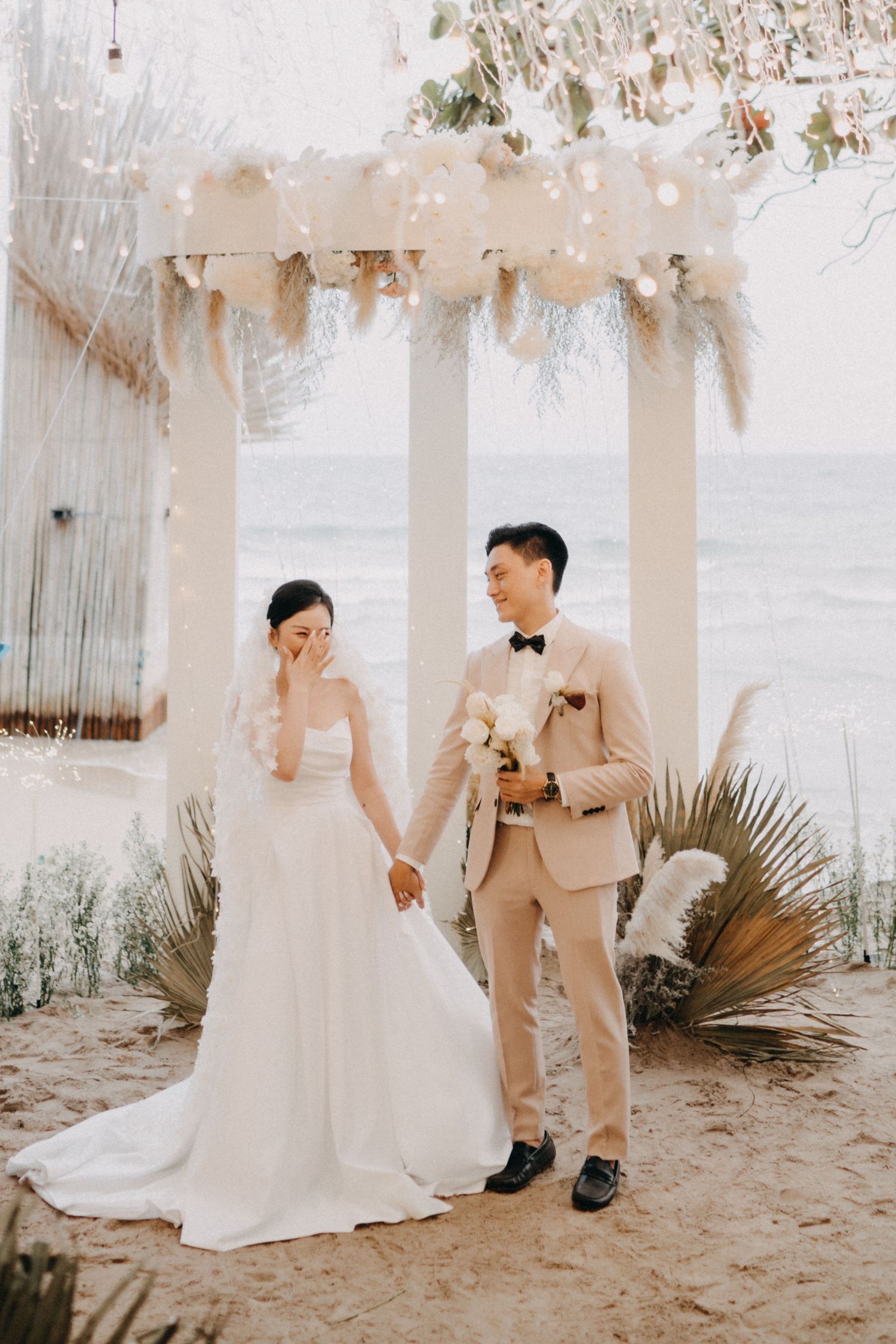 Bride and groom hold hands at a beach wedding ceremony under a floral arch with string lights.