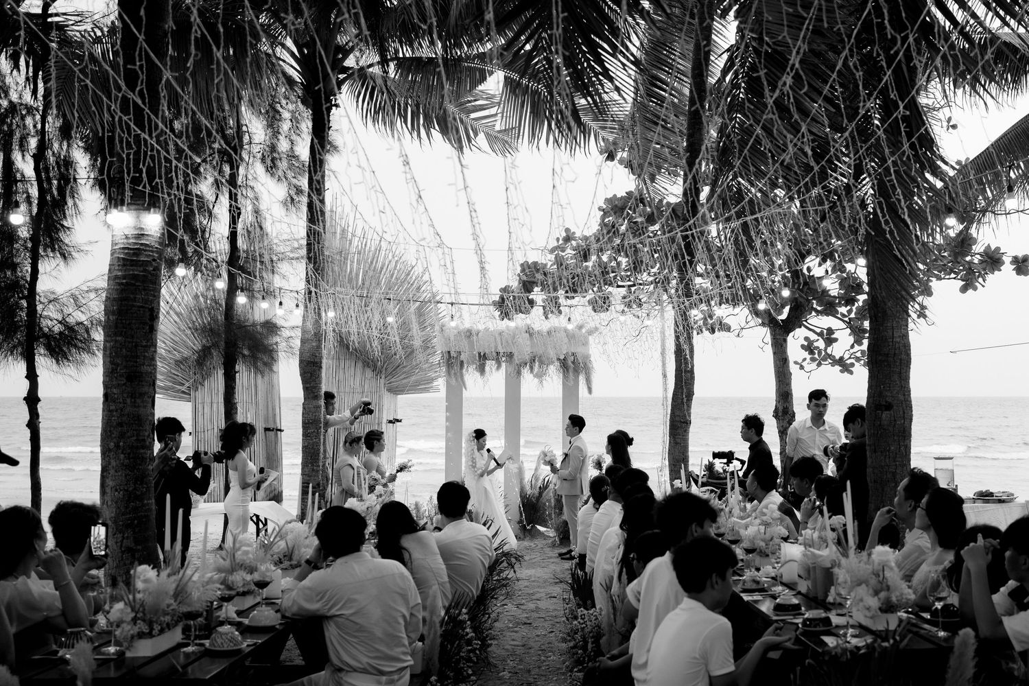 Black and white aerial view of a beach wedding reception under palm trees by the ocean.