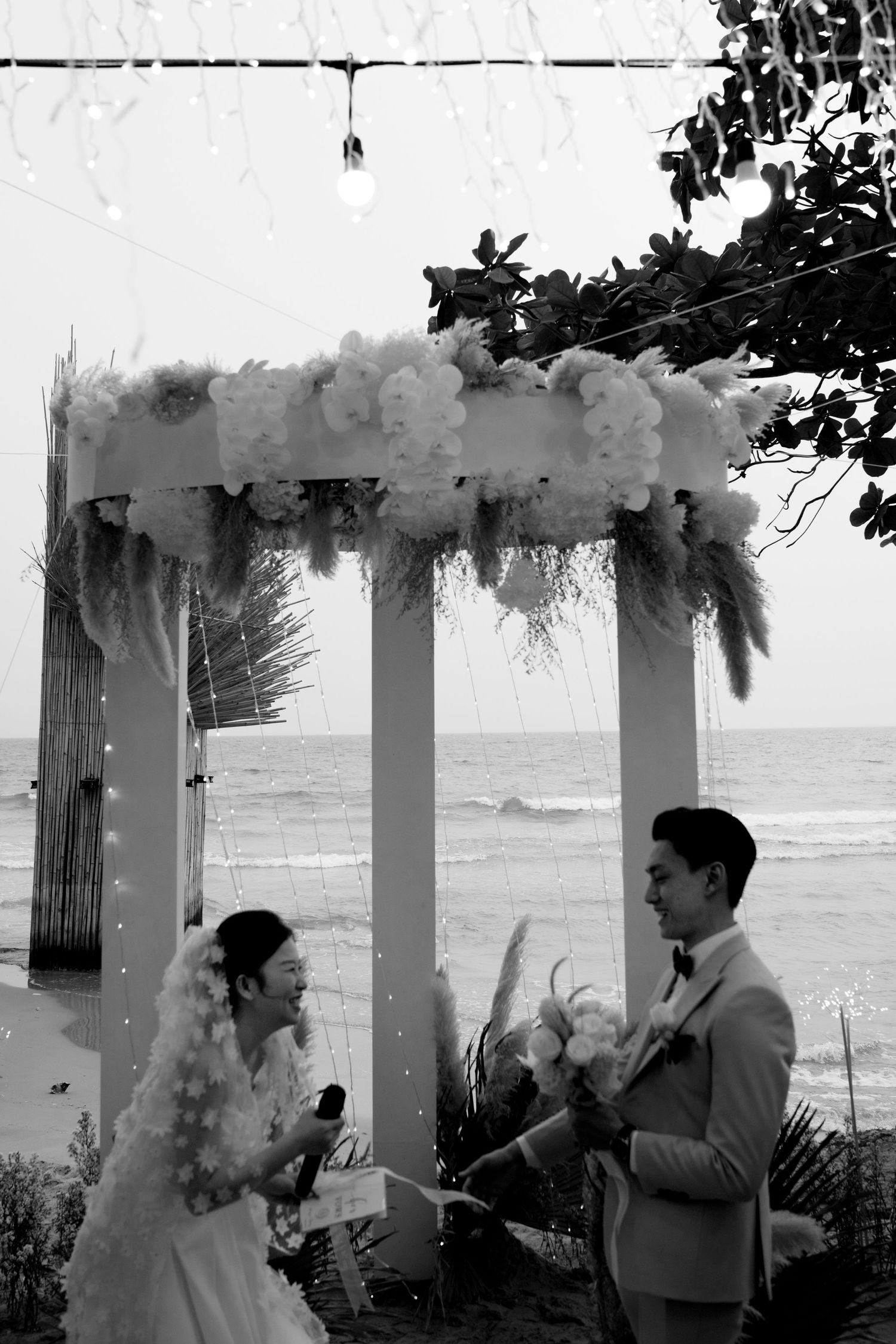 Black and white photo of bride and groom exchanging vows under a floral arch by the sea.
