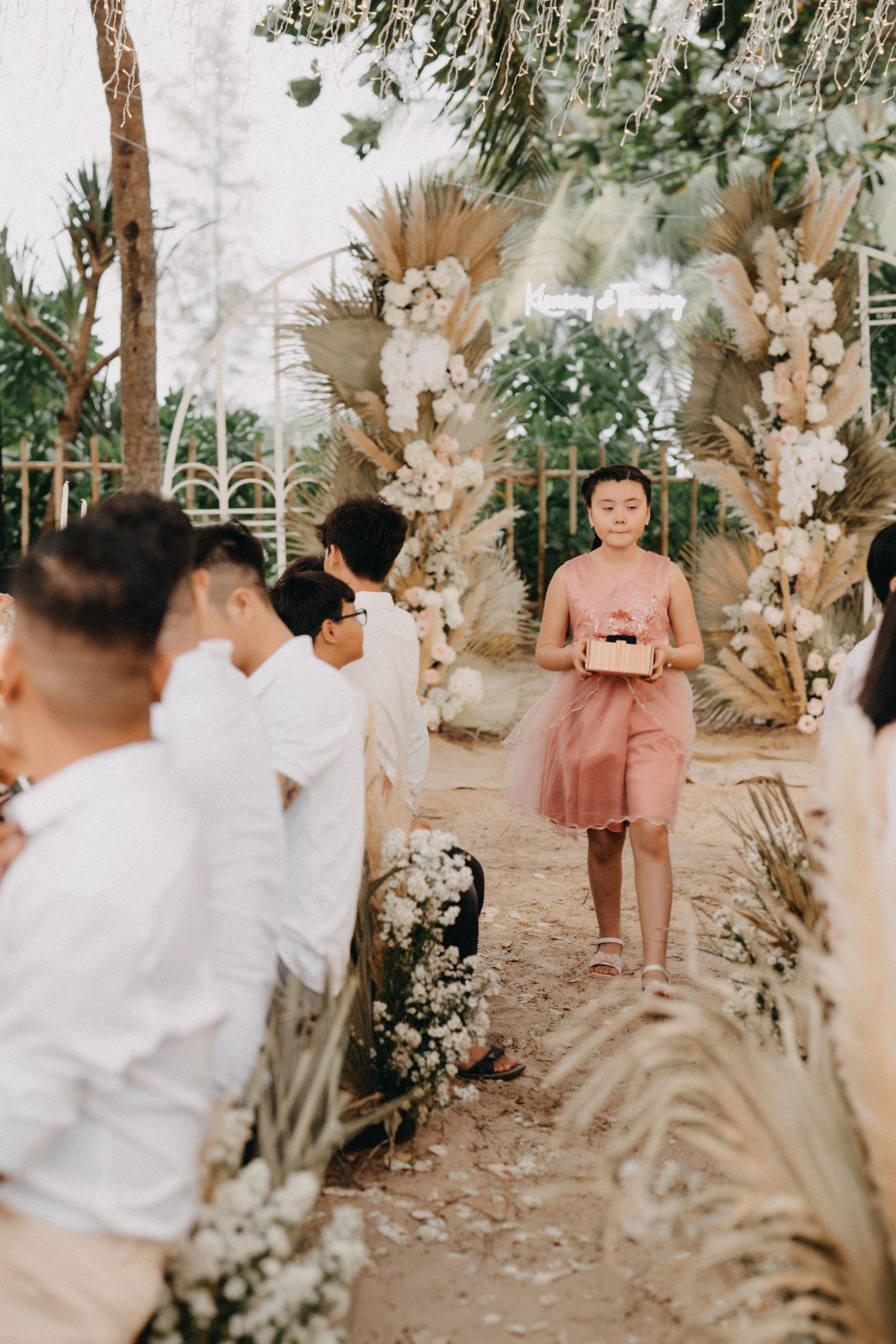Young girl in pink dress walks down sandy wedding aisle carrying a gift box.