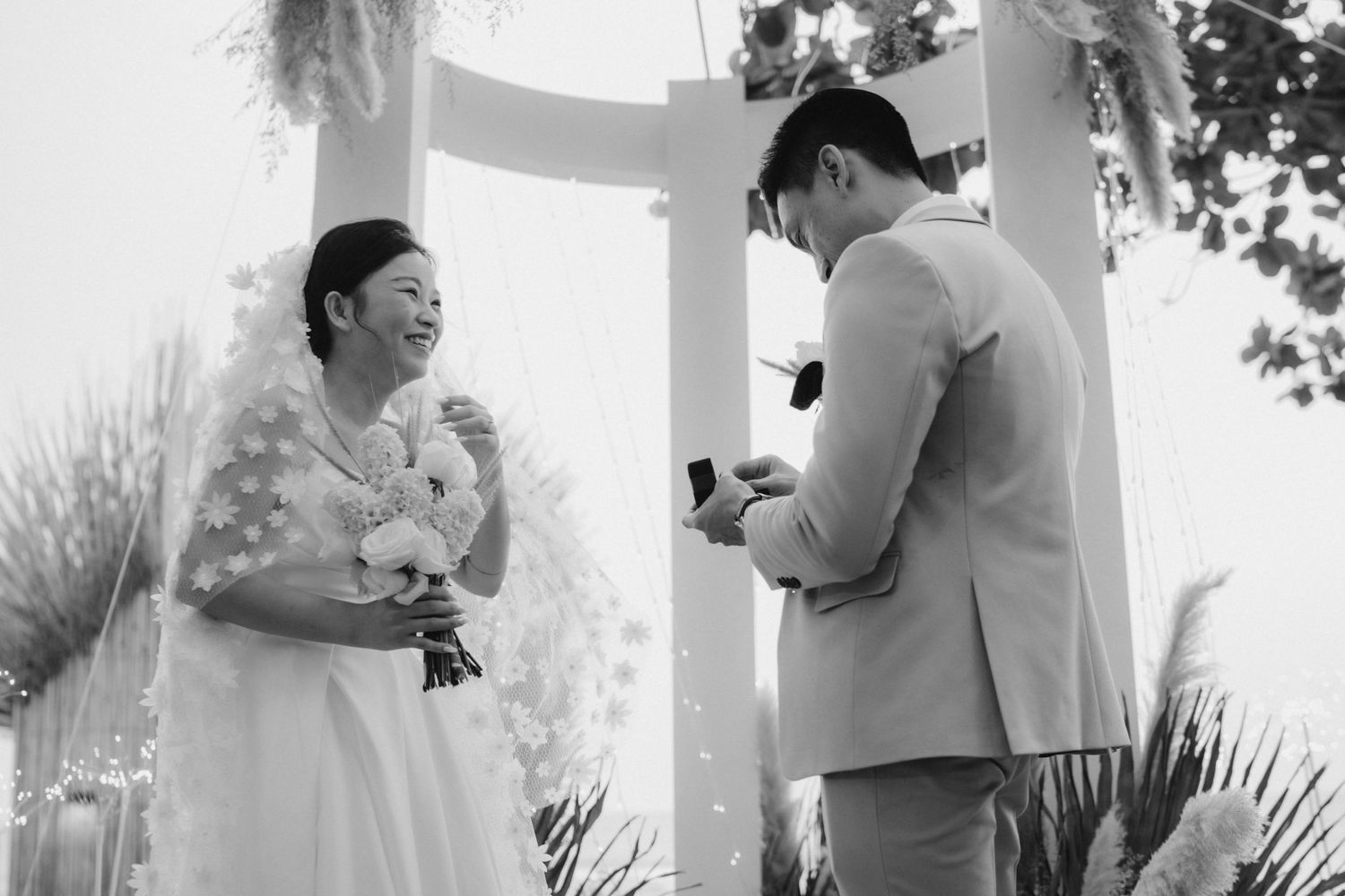 Black and white photo of groom presenting ring to laughing bride at beach ceremony.