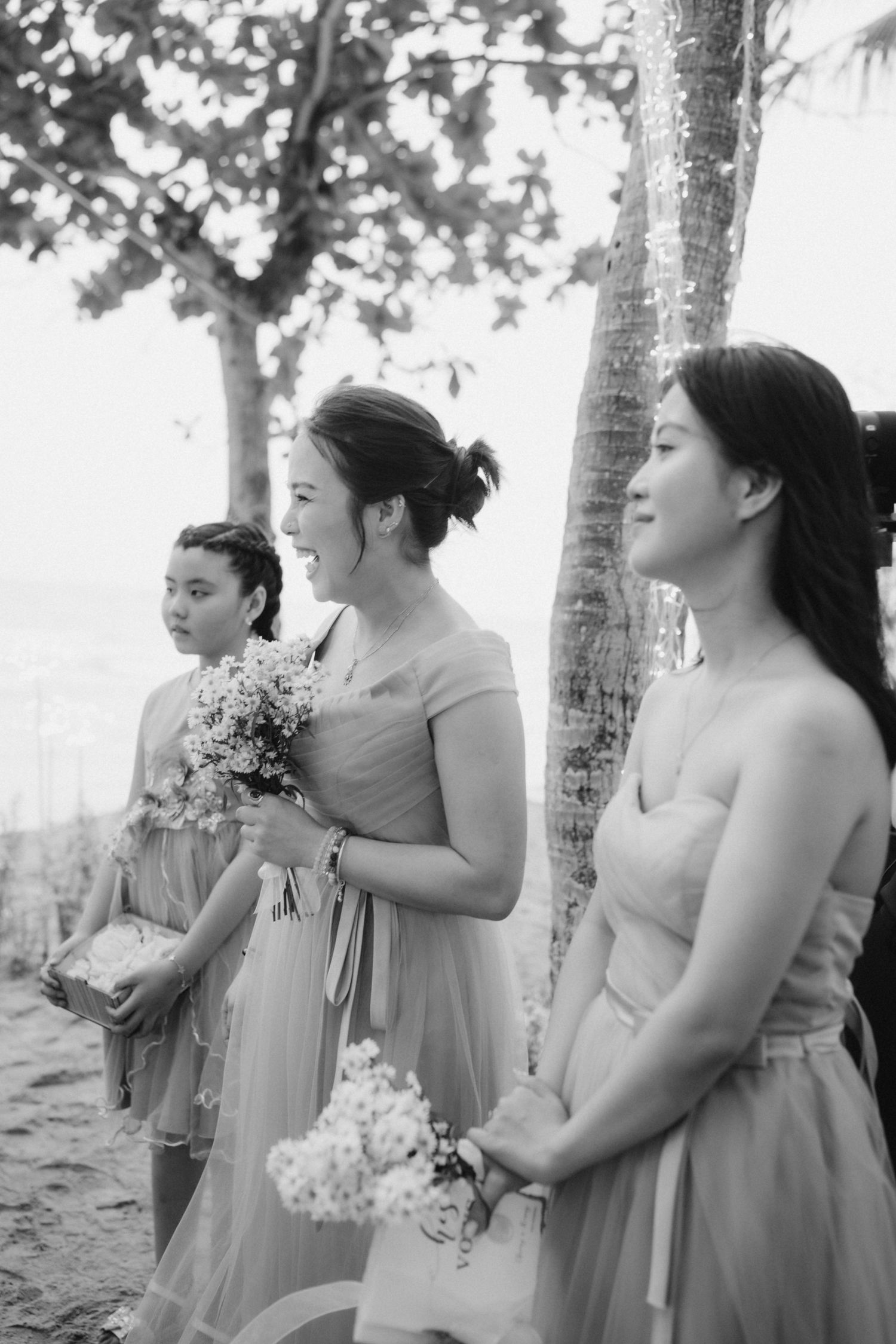 Black and white photo of three bridesmaids in grey dresses holding bouquets by a tree.