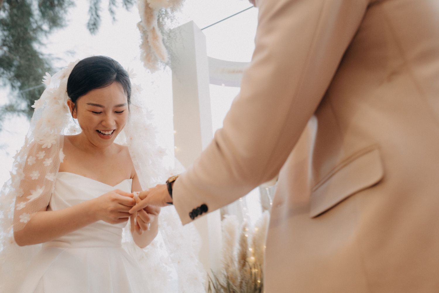 Smiling bride in veil receives wedding ring from groom during indoor ceremony.