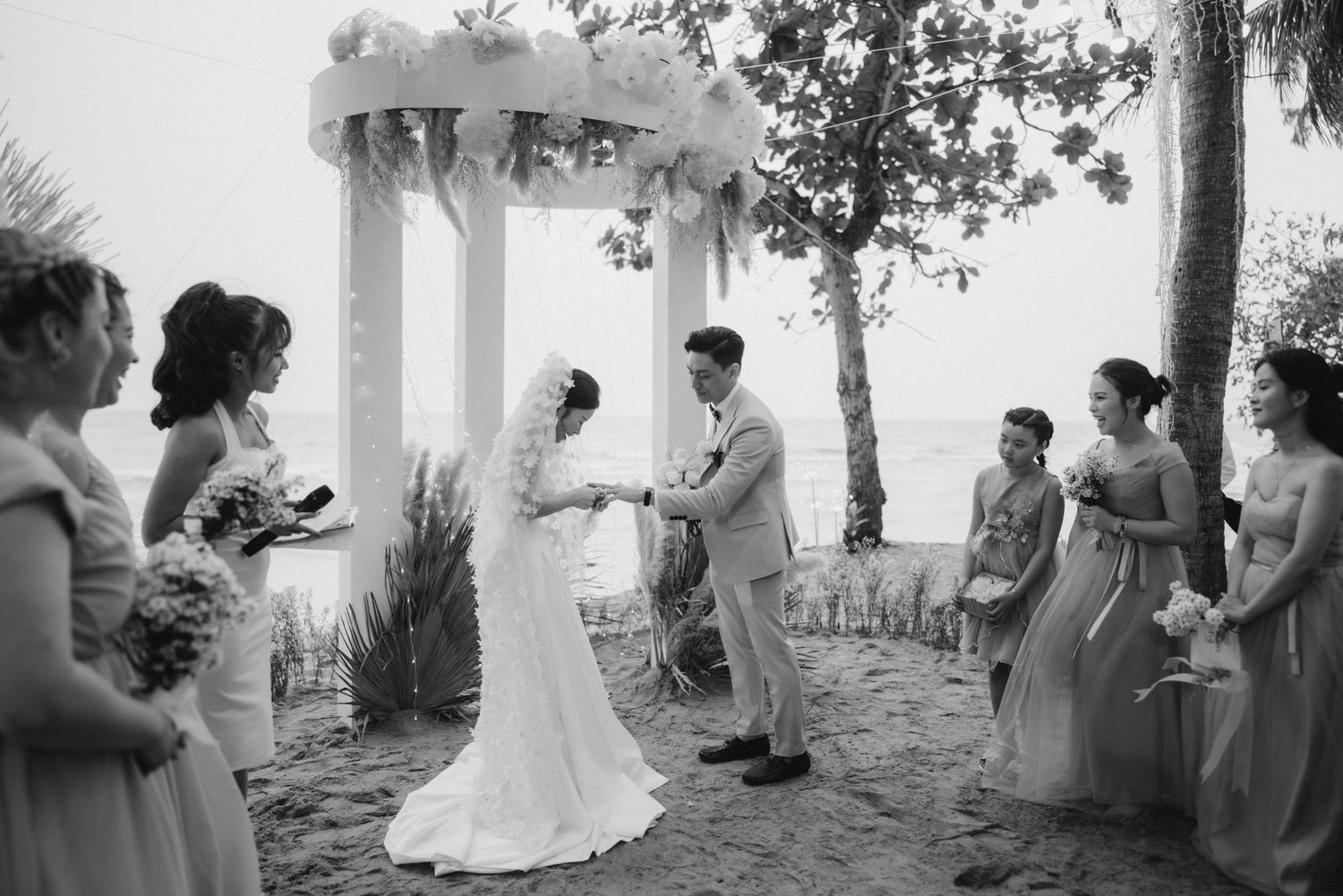 Black and white photo of bride and groom holding hands at beach altar with wedding party.