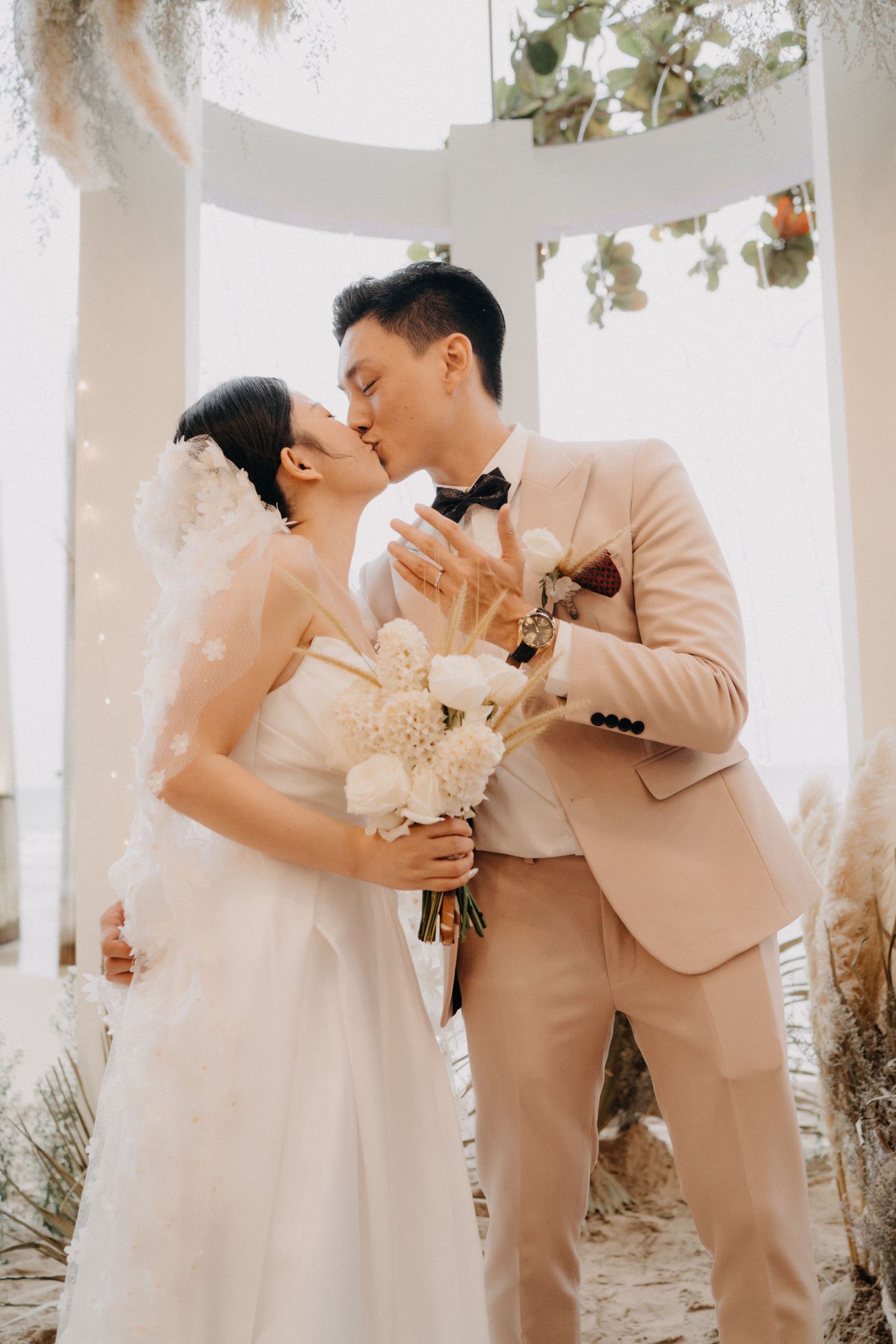 Bride and groom share a kiss under a floral arch at their beach wedding ceremony.