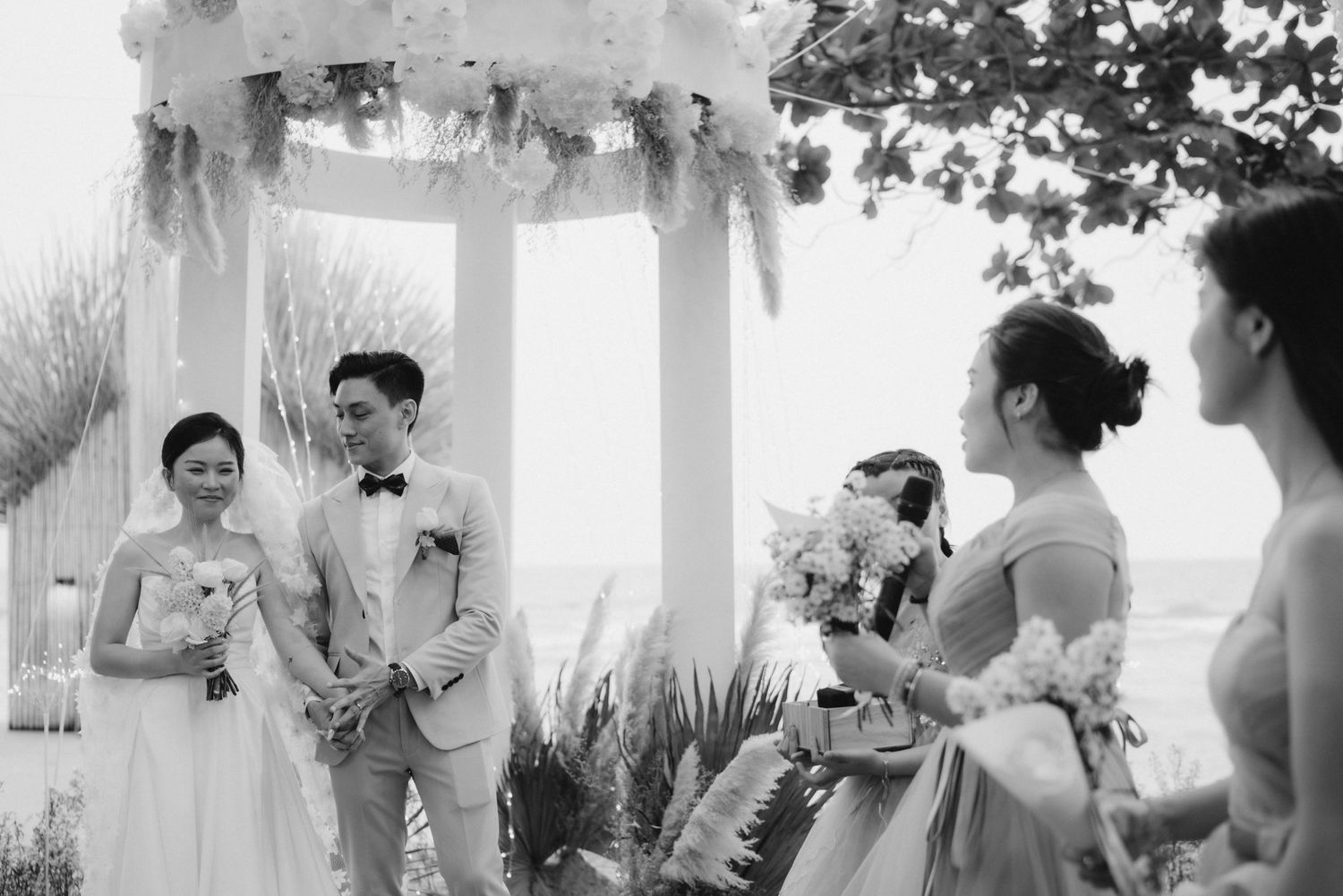 Bride and groom stand at outdoor wedding altar with bridesmaids and guests in black and white photo.