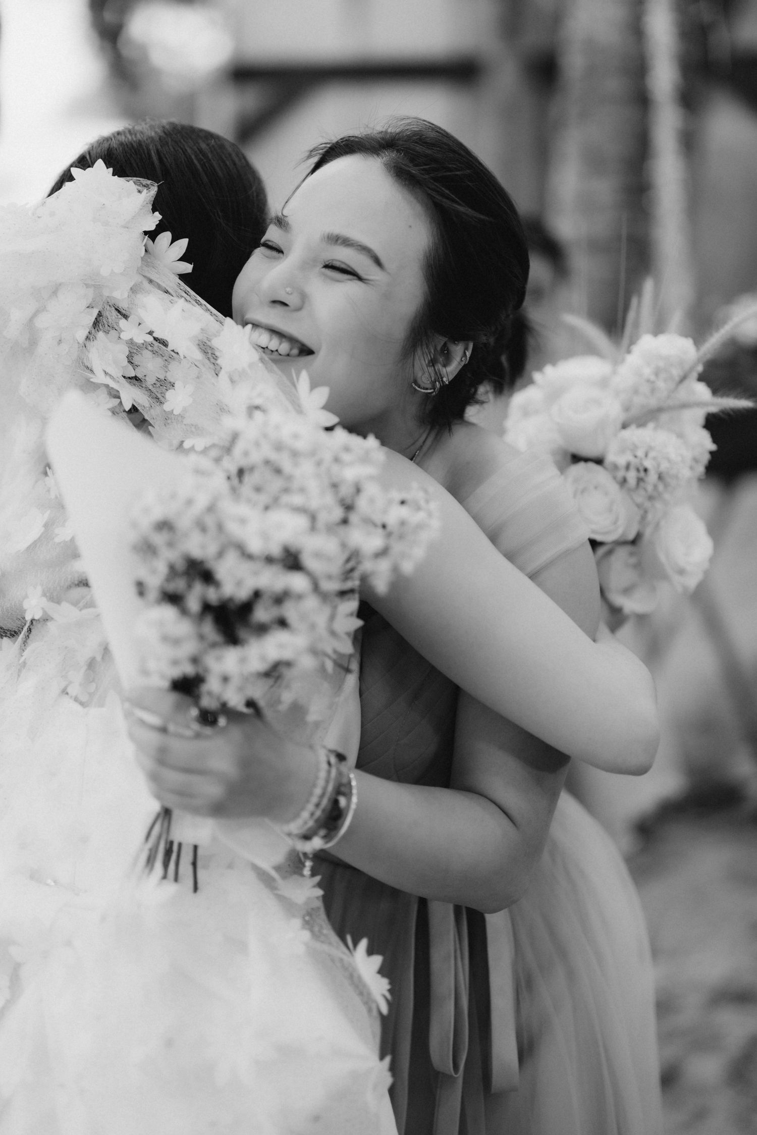 Joyful bridesmaid hugs bride holding floral bouquet in black and white wedding photo.