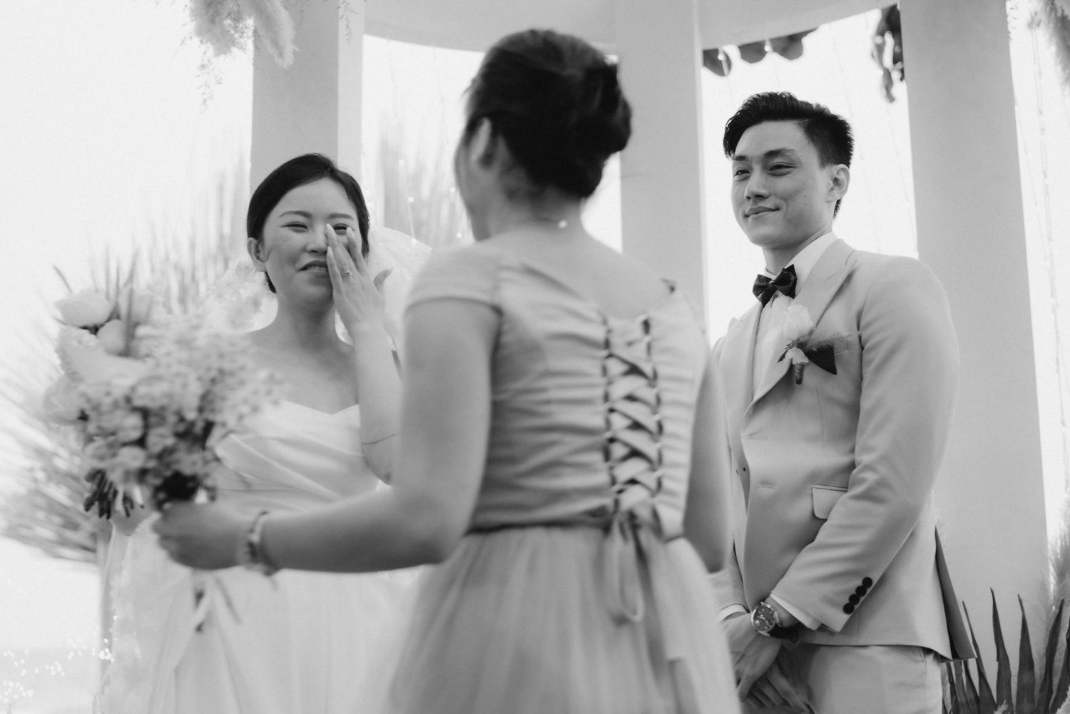 Emotional bride wipes tear as groom smiles during outdoor wedding ceremony in black and white.