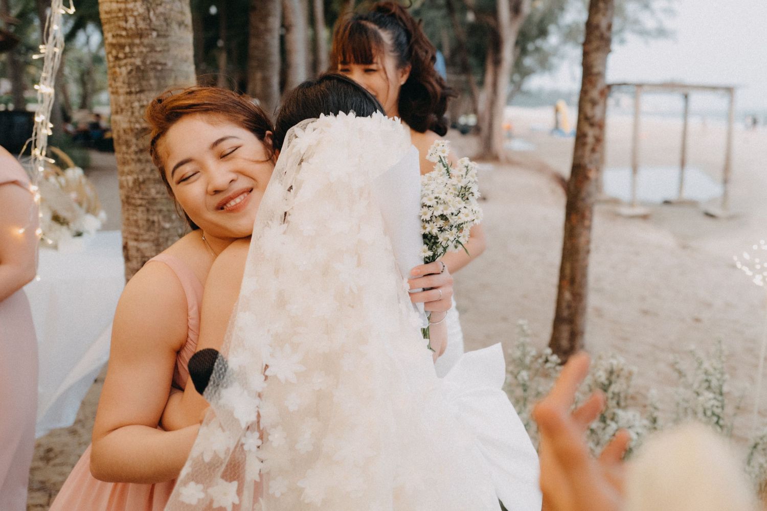 Bridesmaid in pink dress embraces bride in floral veil near palm trees at beach wedding.