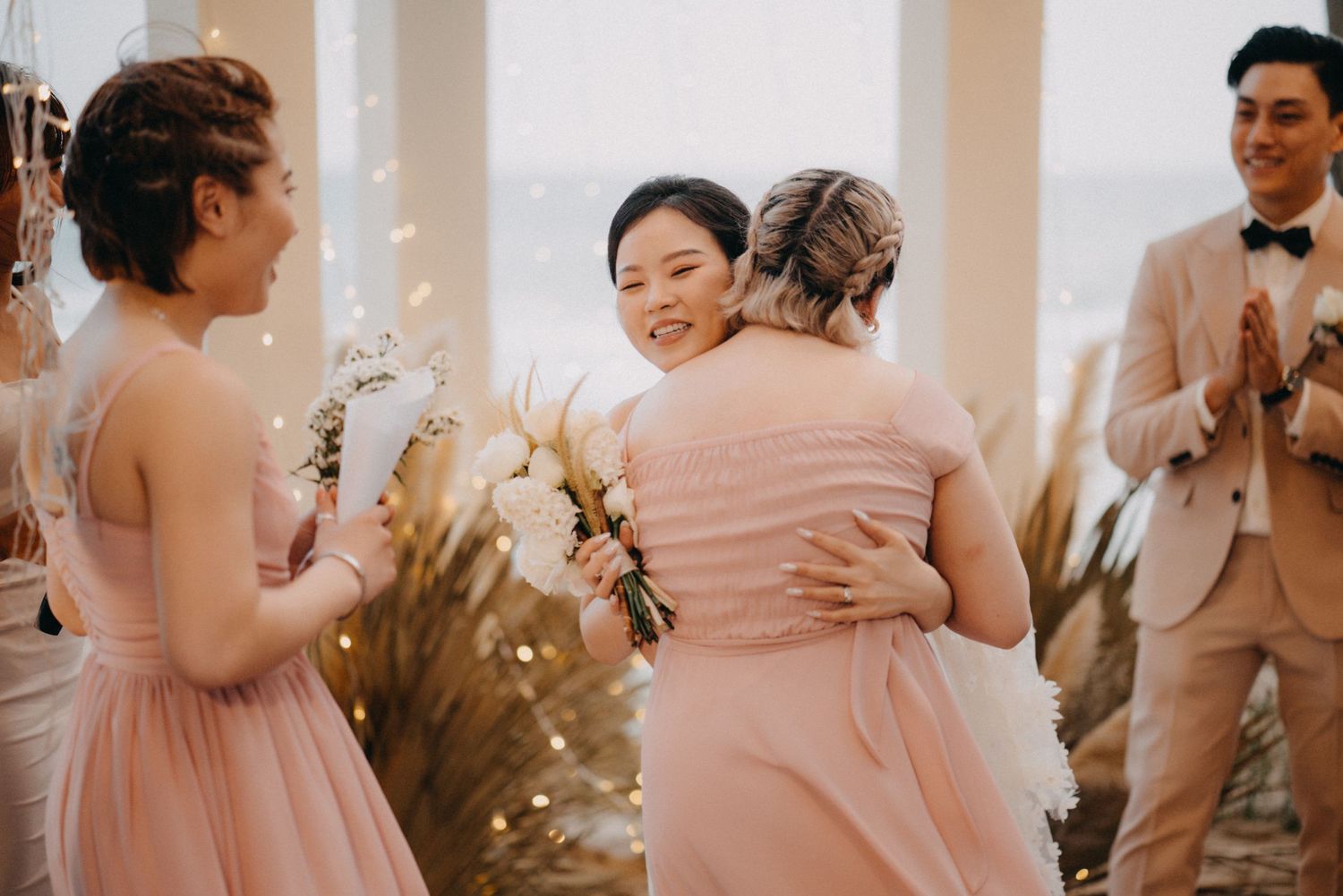 Bride hugs bridesmaid in pink dress while groom applauds at elegant indoor wedding ceremony.