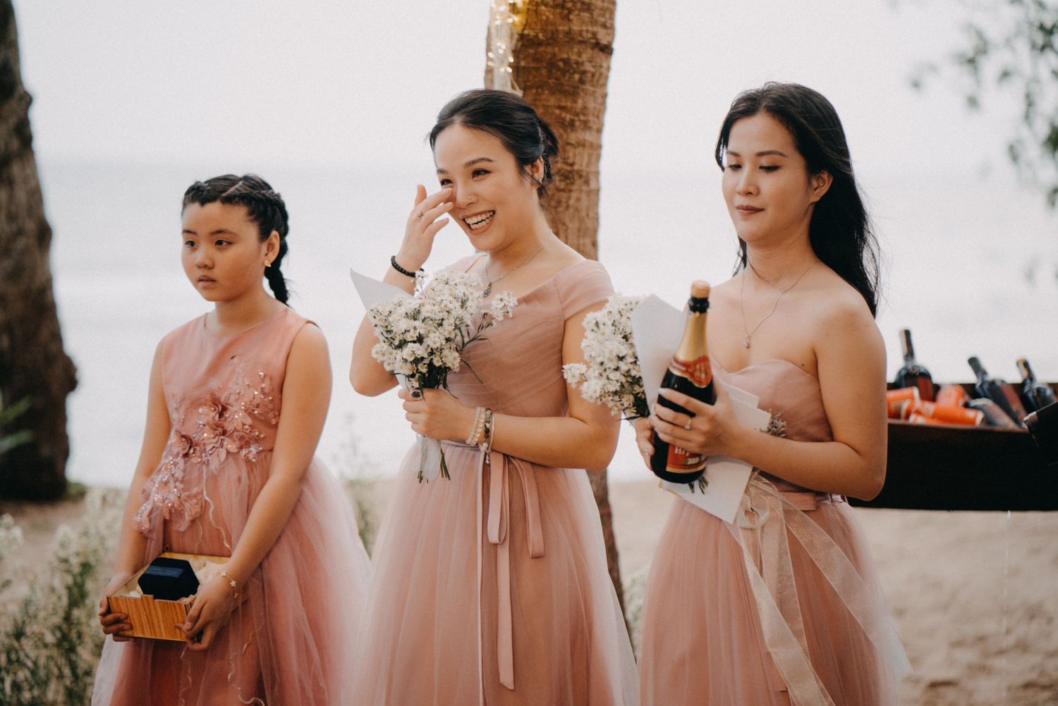 Three bridesmaids in blush pink dresses hold bouquets and champagne at beachside wedding celebration.