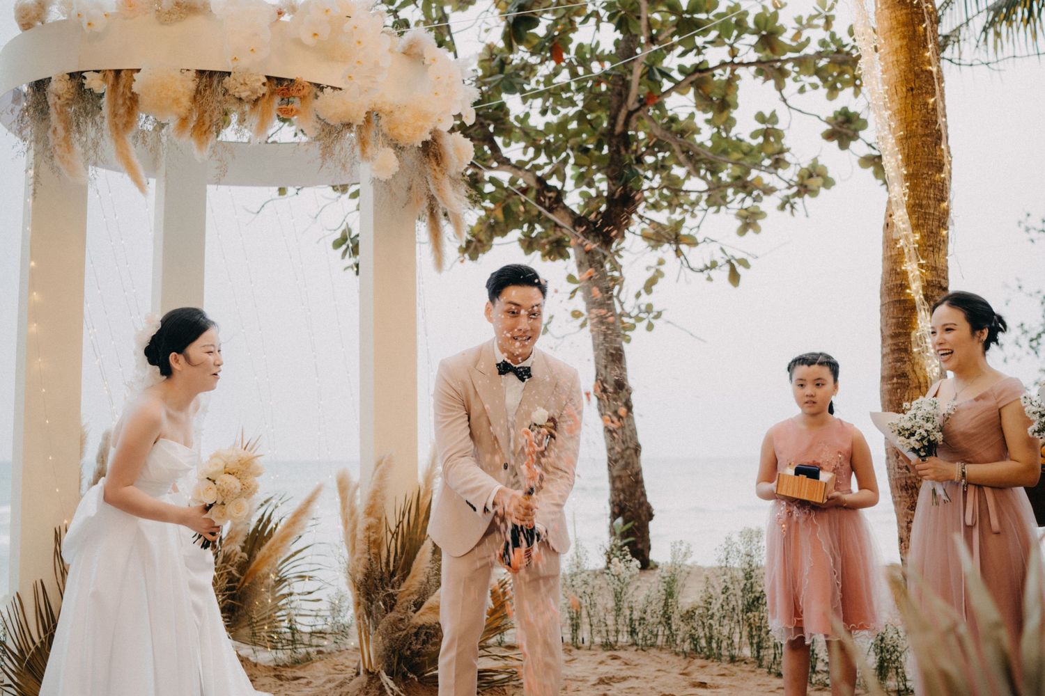 Bride and groom celebrate at beach wedding ceremony with bridesmaids under floral arch.