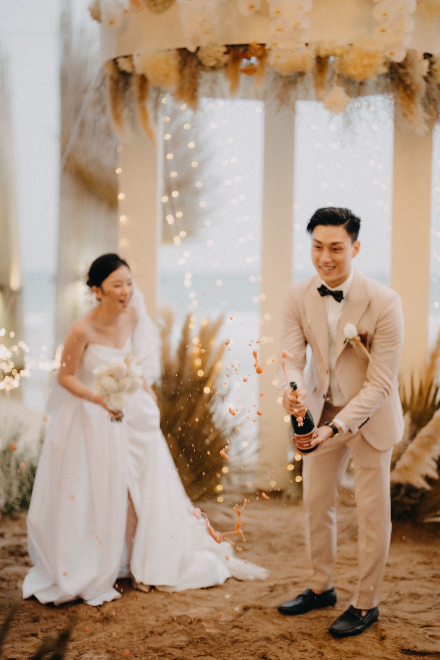 Groom pops champagne bottle at elegant beach wedding with string lights and floral decor.