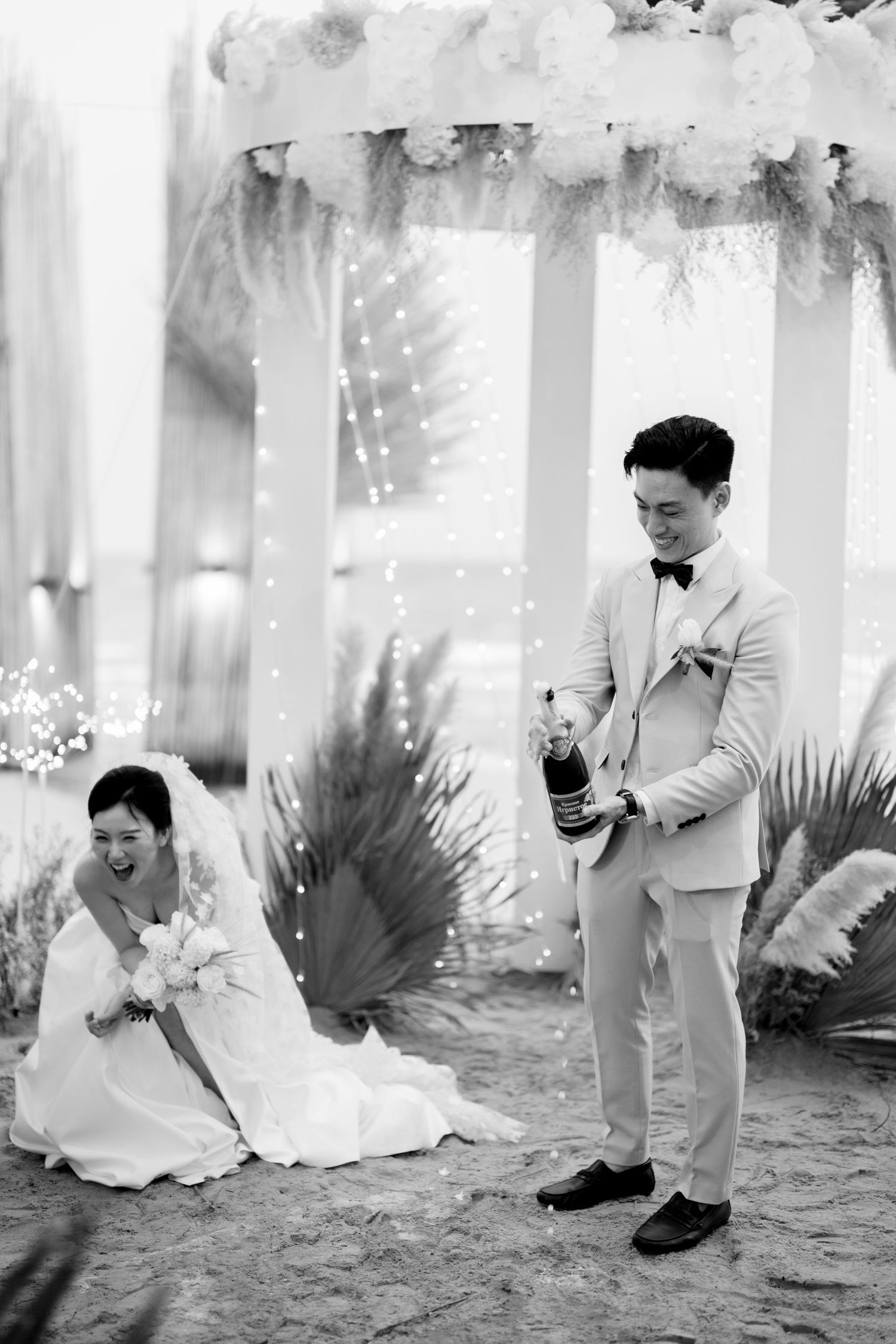 Black and white photo of groom popping champagne as bride laughs at beach wedding.