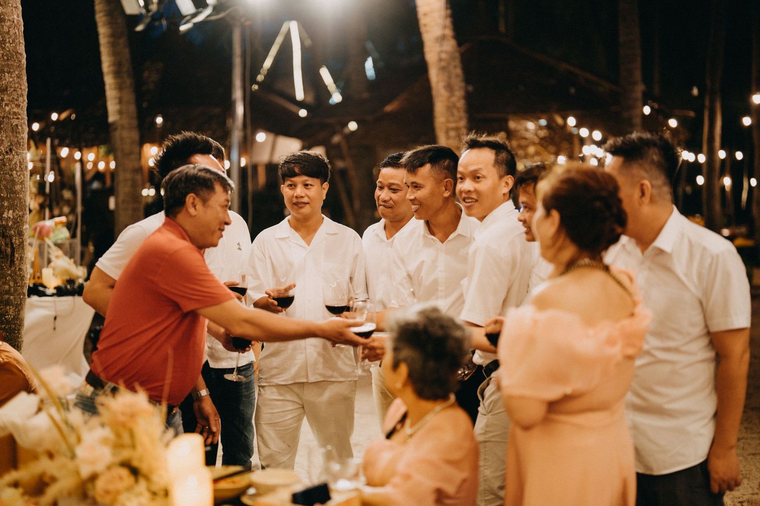 Wedding guests in white shirts toast and celebrate at outdoor beach reception at night.