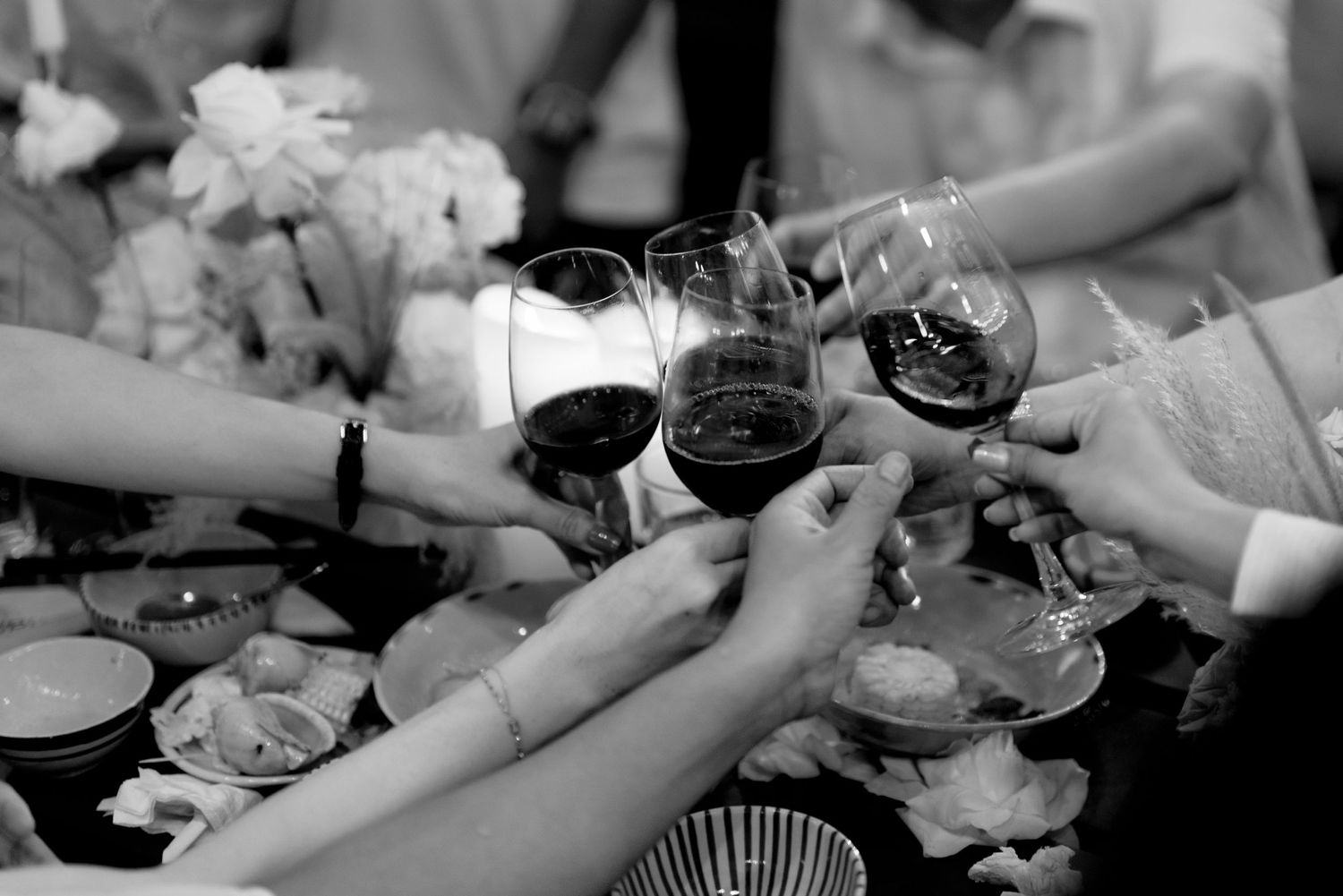 Black and white close-up of wedding guests clinking red wine glasses at reception dinner.