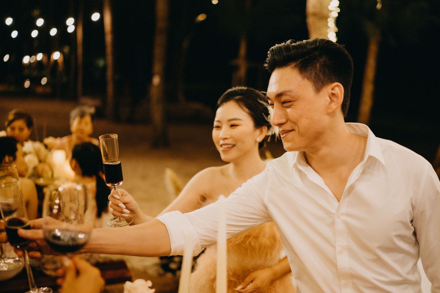 Bride and groom raise red wine glasses for a toast at romantic outdoor wedding reception.