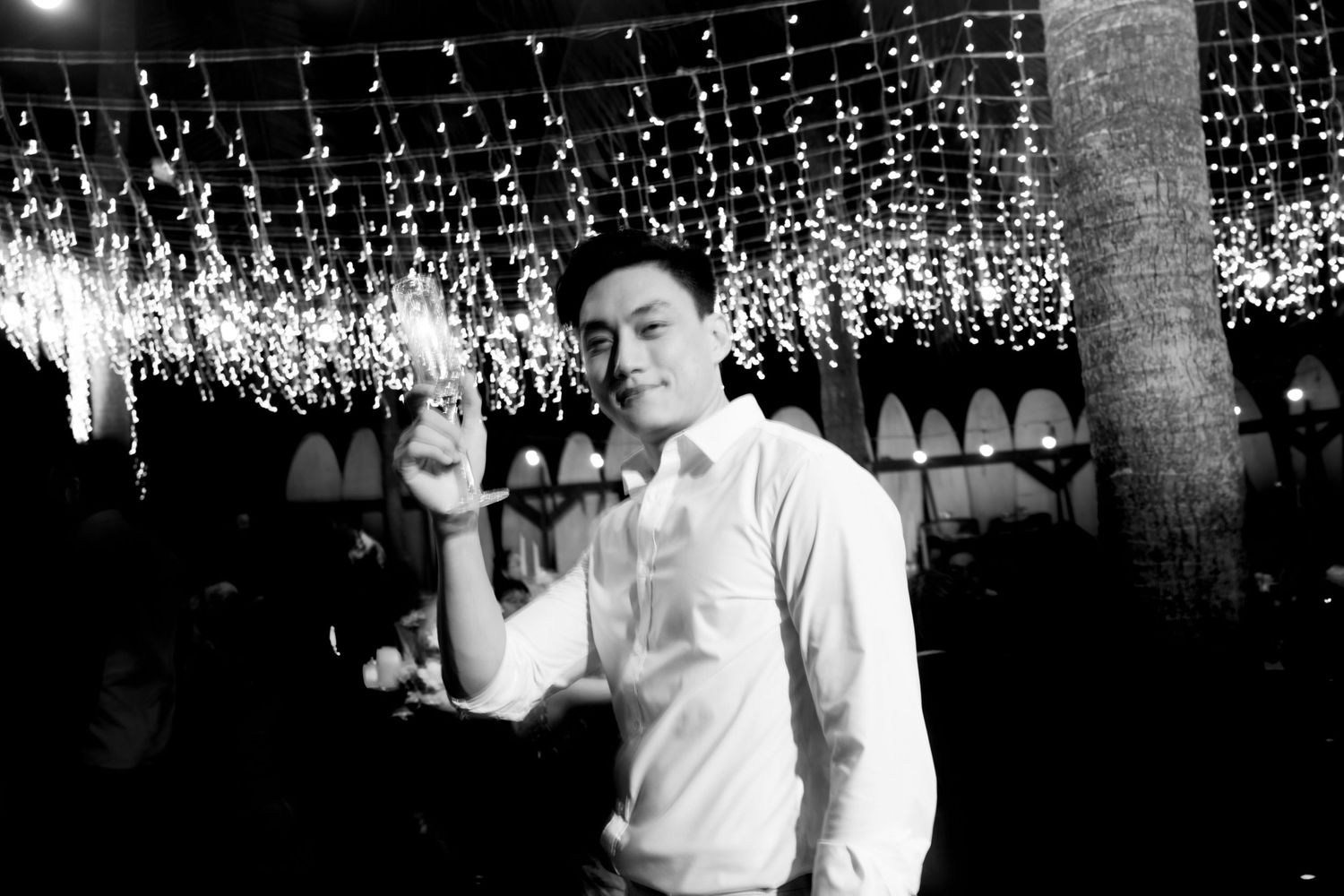Young man in white shirt pointing finger up at elegant outdoor night event with fairy lights.
