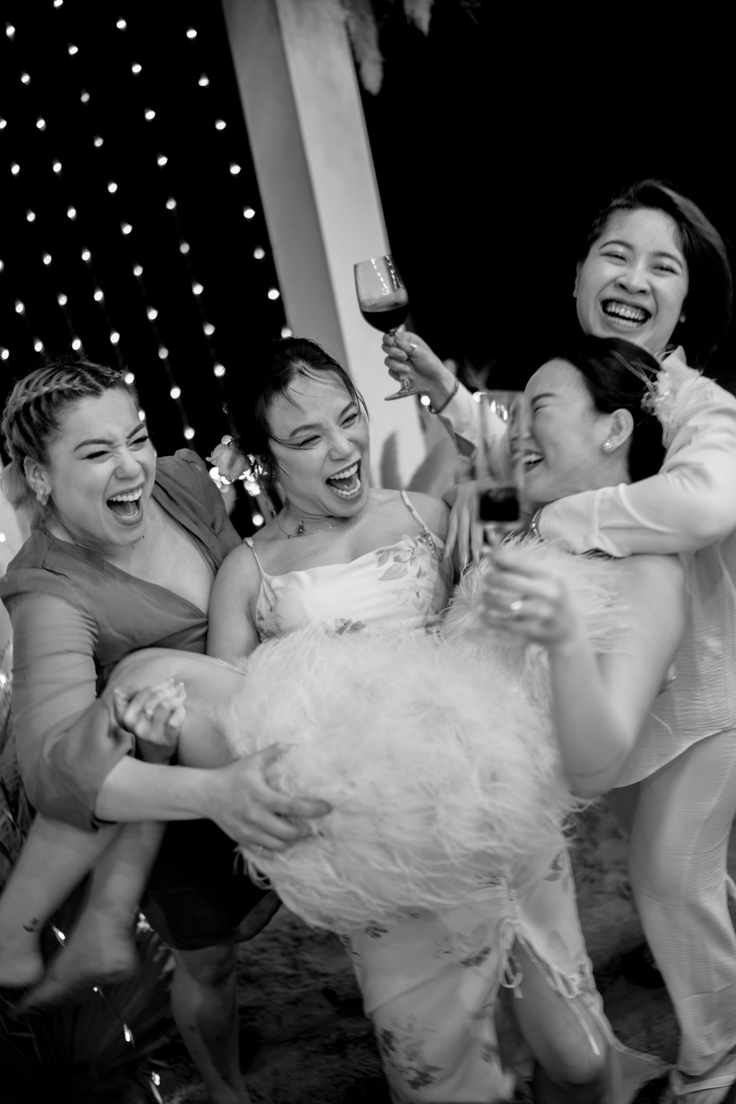 Bridesmaids joyfully lifting bride in wedding dress while laughing at outdoor reception.