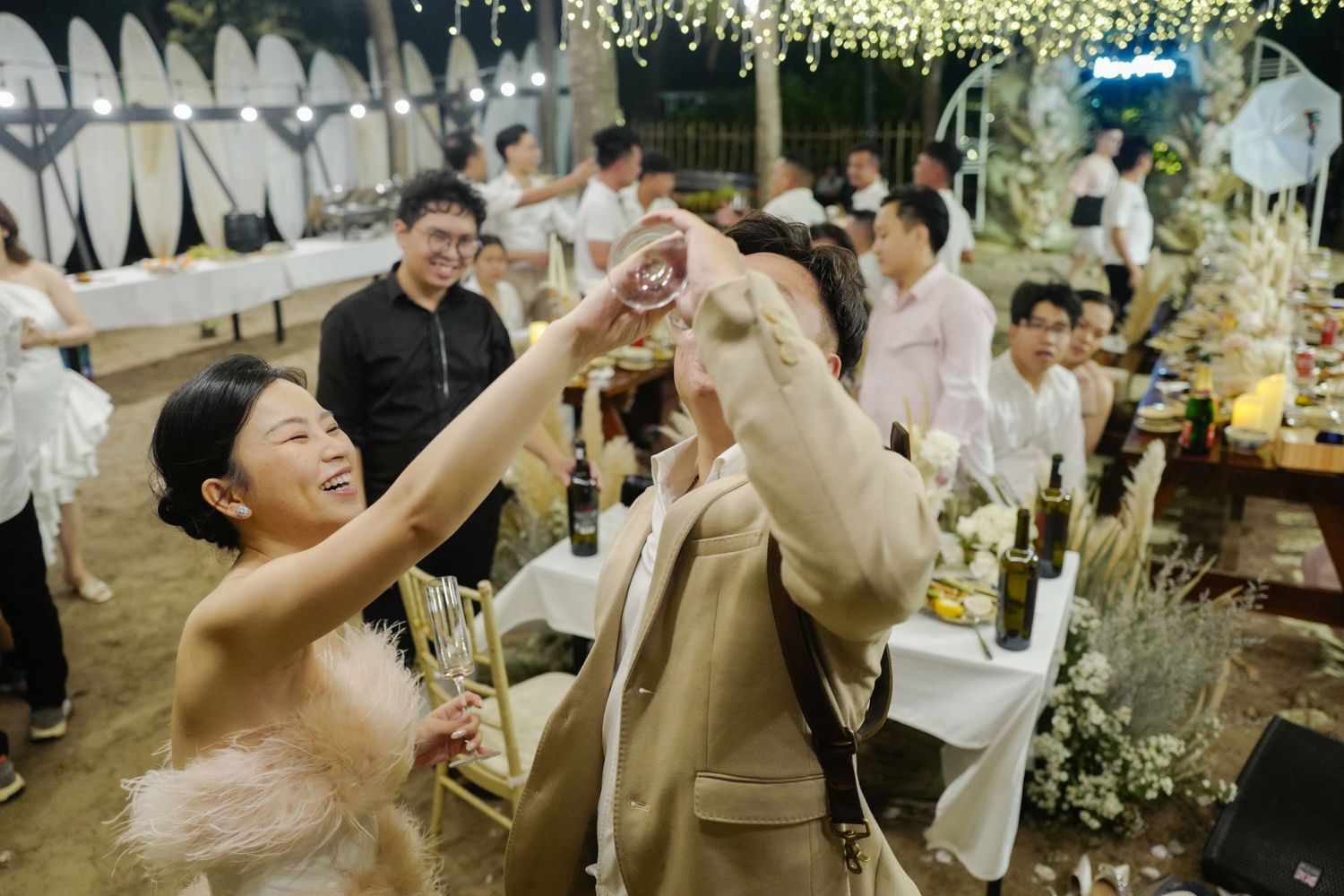 Bride in pink dress toasting drinks with guest at decorated outdoor wedding reception.