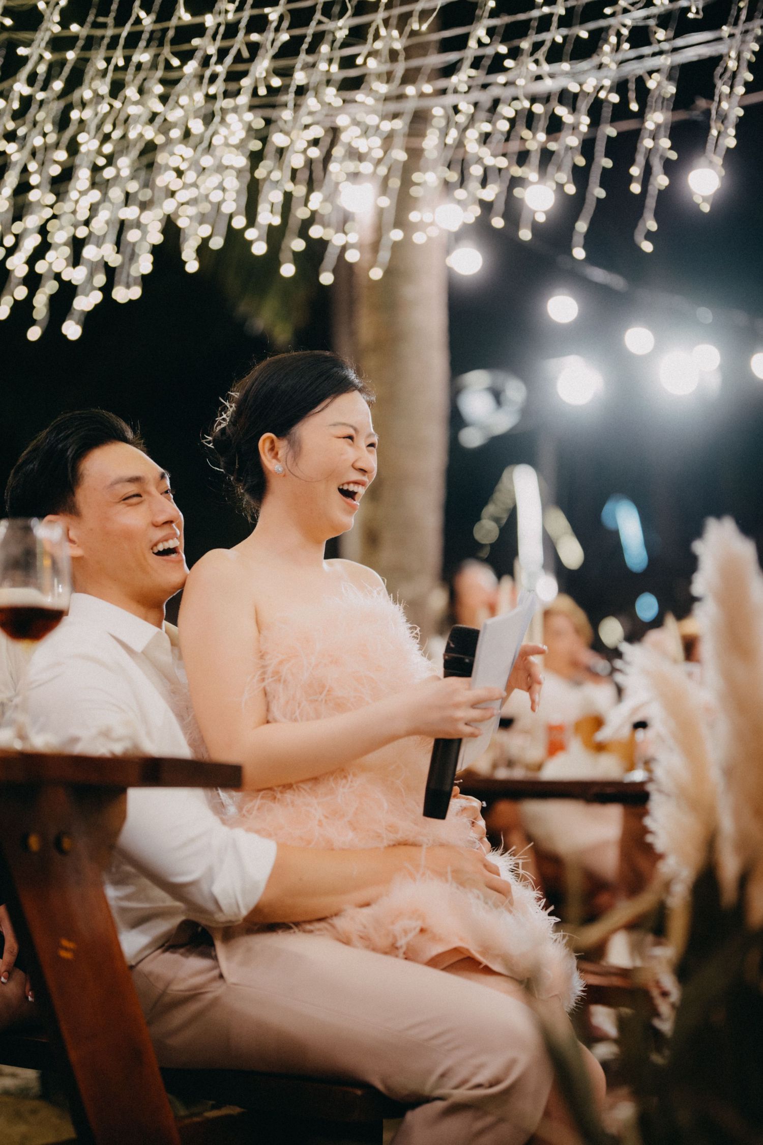 Smiling bride and groom sitting together at outdoor wedding reception under string lights.