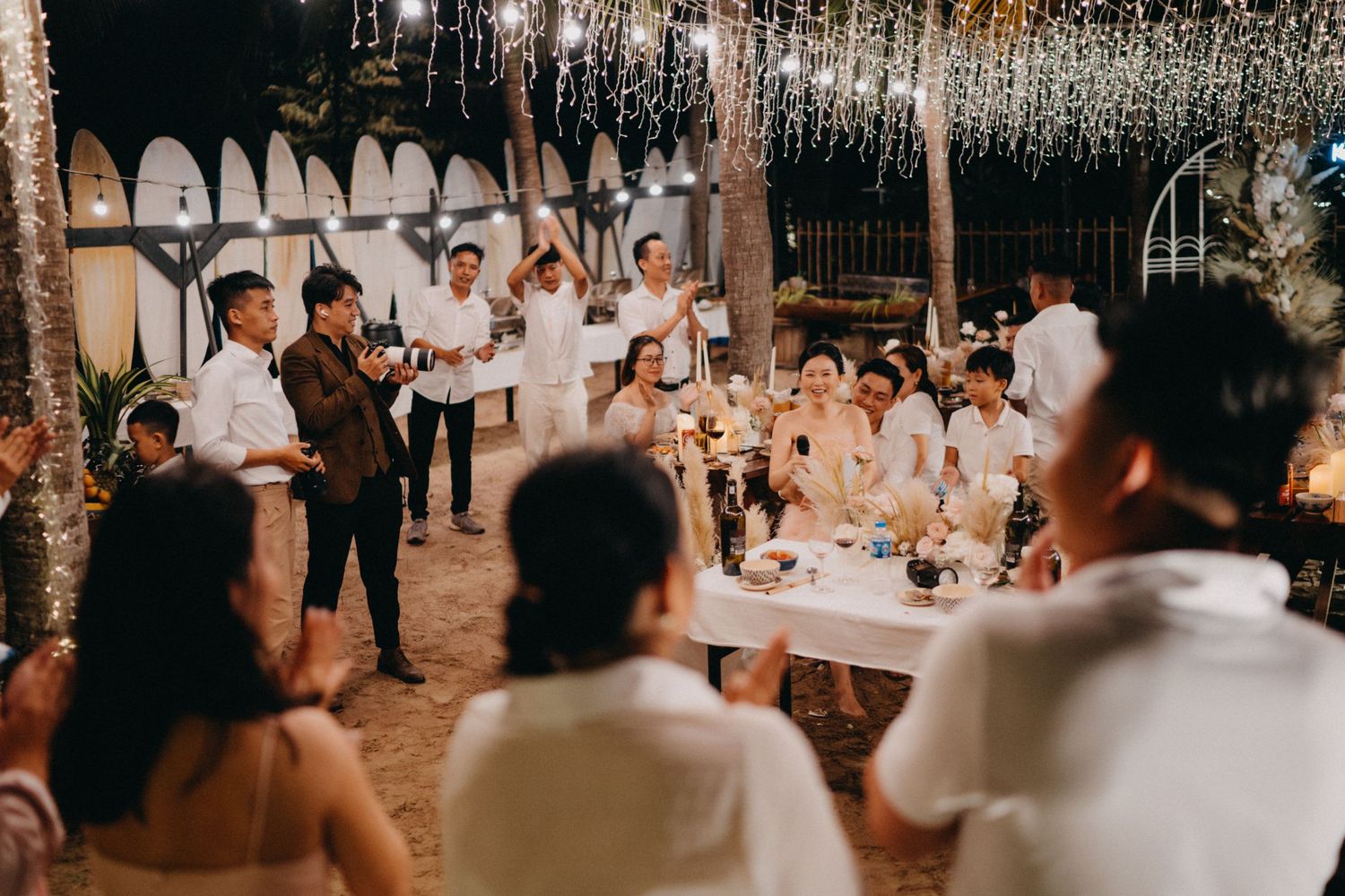 Wedding guests gathered around decorated table at elegant outdoor evening reception.