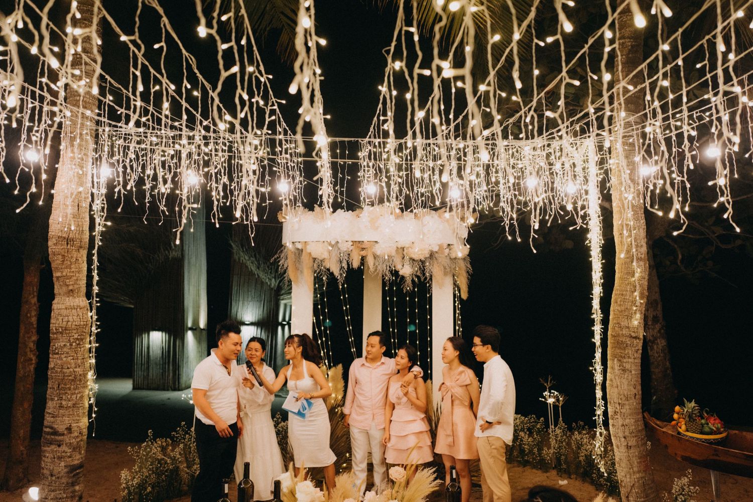 Wedding party group posing under cascading fairy lights at outdoor nighttime celebration.