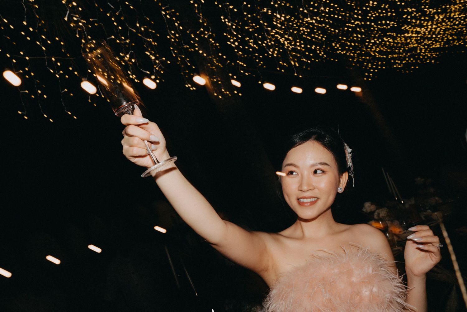 Young Asian woman in pink feather dress raises sparkler under twinkling lights at elegant evening celebration.