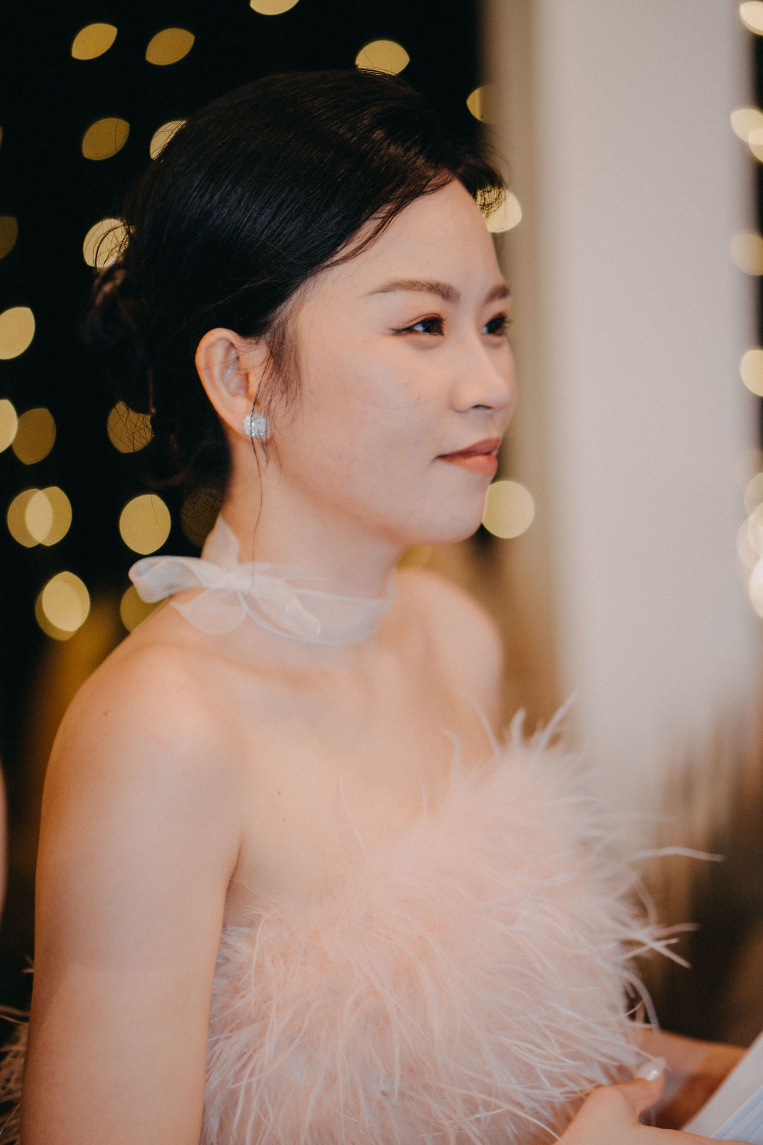 Elegant Asian woman in strapless pink feather dress with blue earrings poses against bokeh light background.