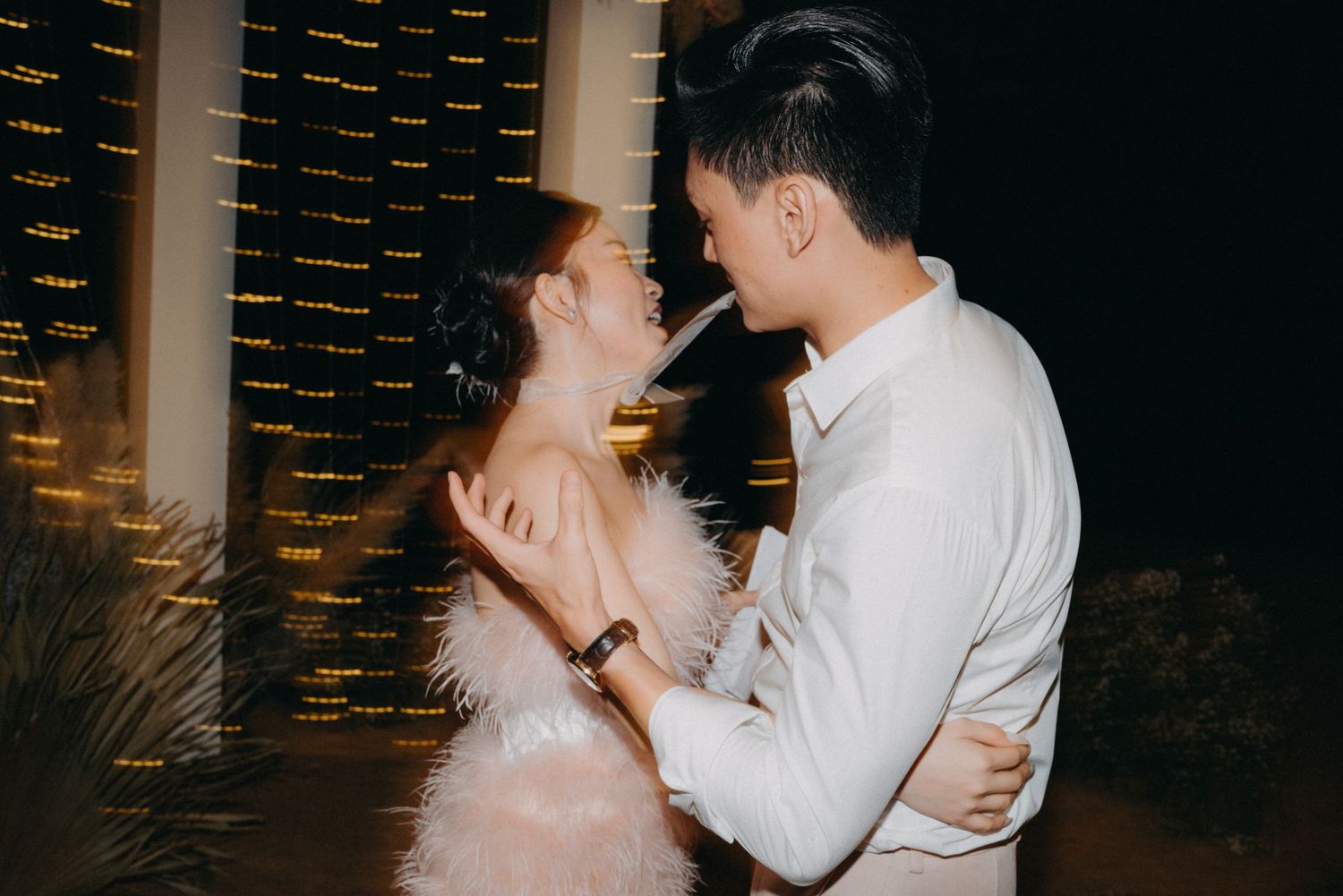 Asian couple sharing romantic moment, woman in pink feather dress and man in white shirt under evening lights.