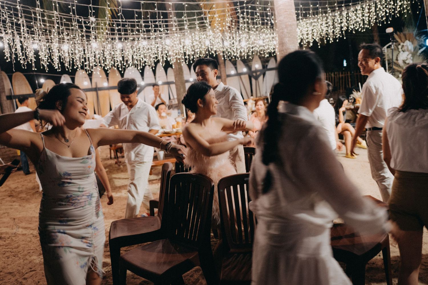 Guests dancing joyfully under hanging floral lights at an outdoor wedding reception on sandy venue.