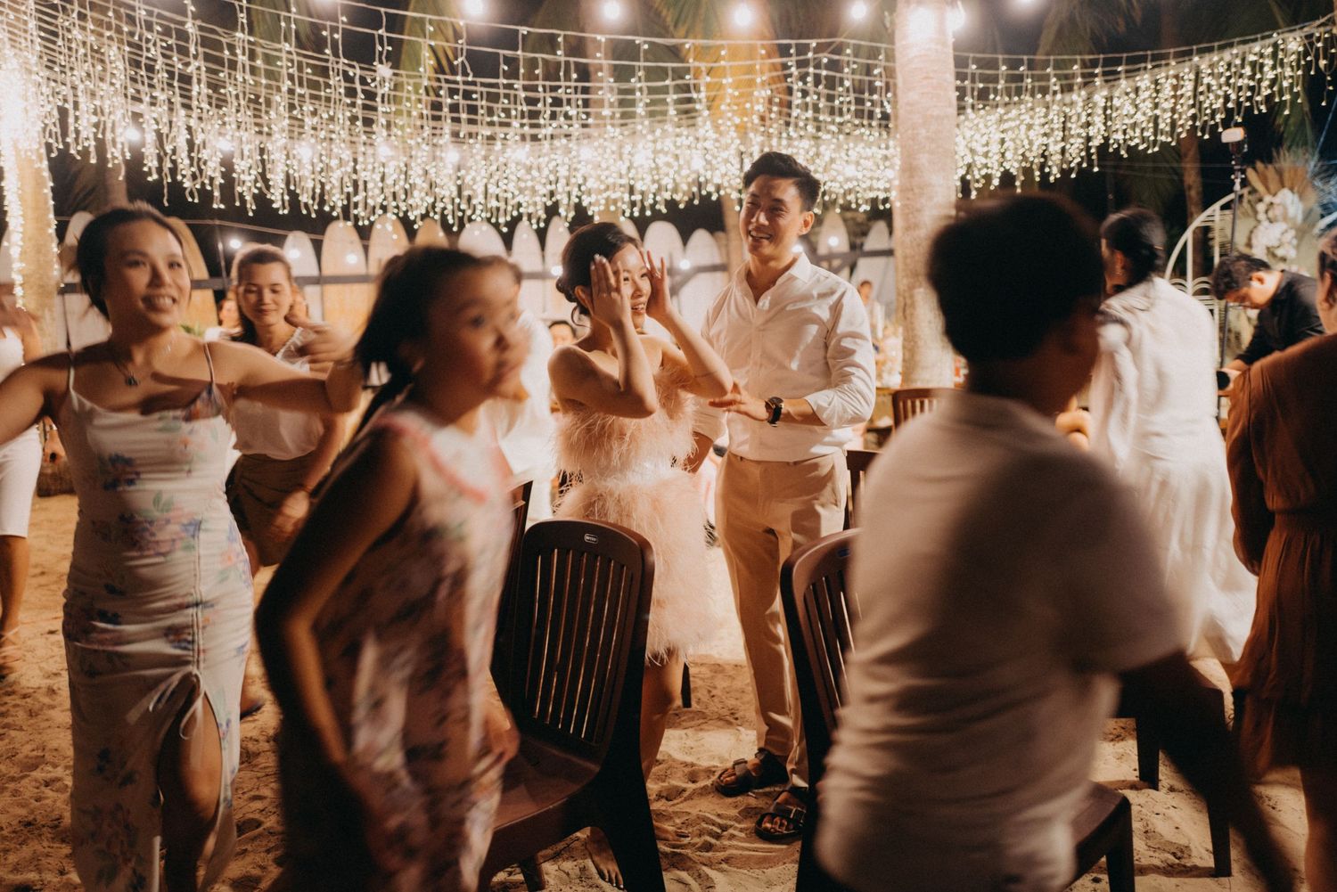 Wedding guests dancing under string lights at a warm outdoor beach reception with floral decorations.