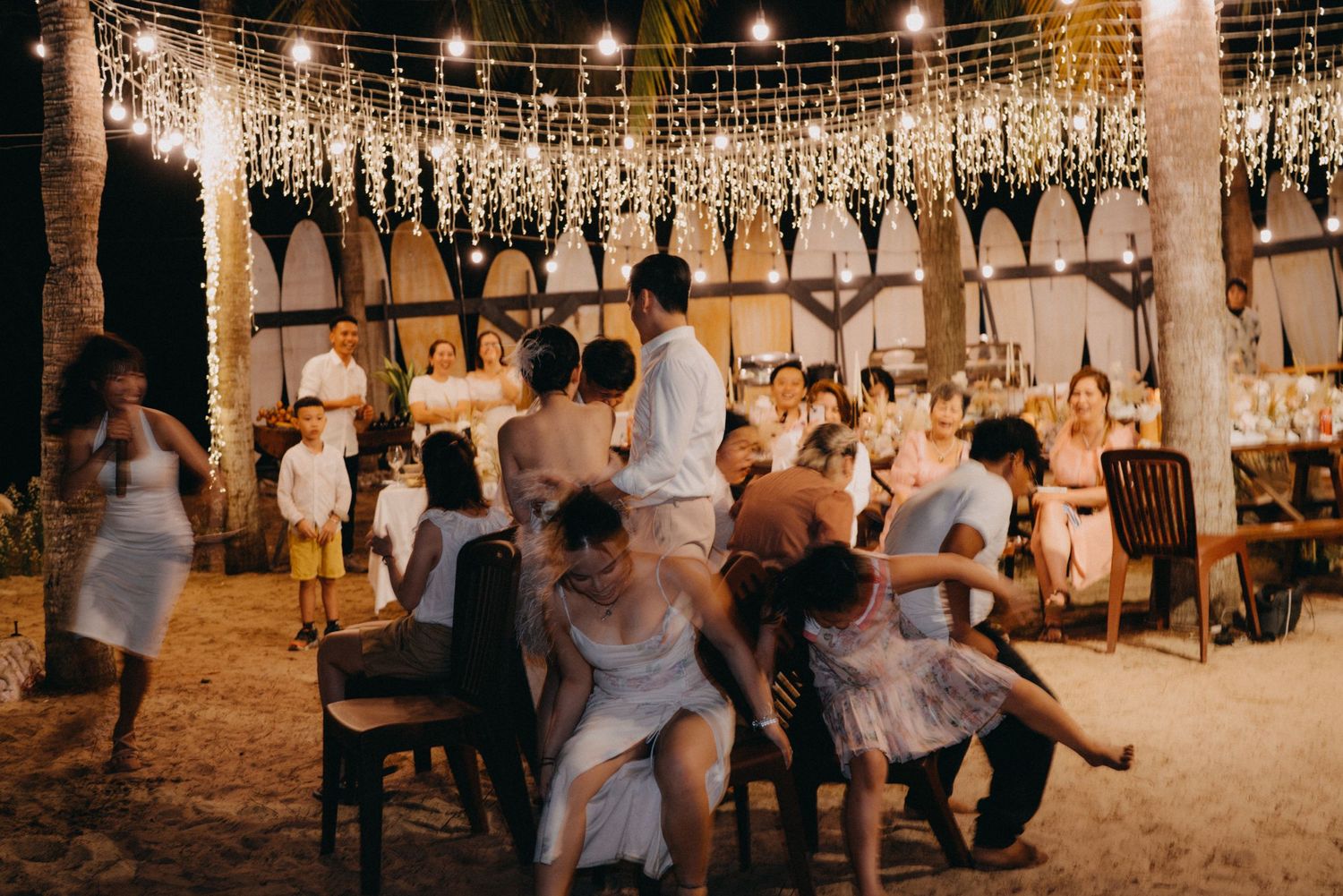 Wide shot of lively wedding reception with guests dancing under fairy lights at outdoor beach venue.