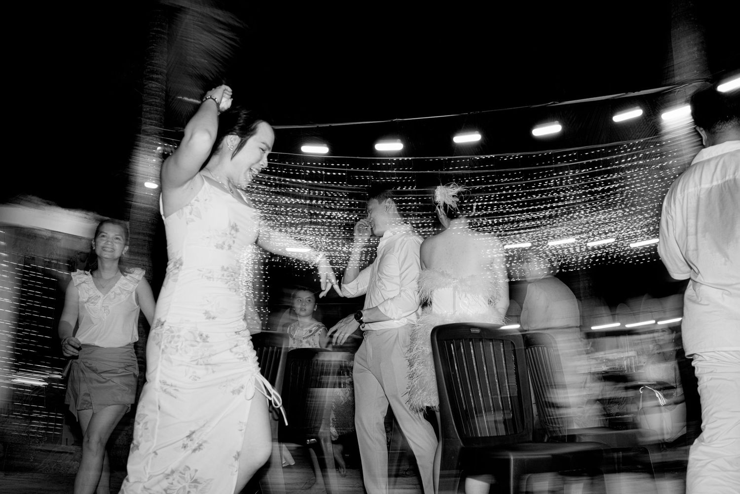 Black and white photo of wedding guests dancing joyfully under string lights at an outdoor evening reception.