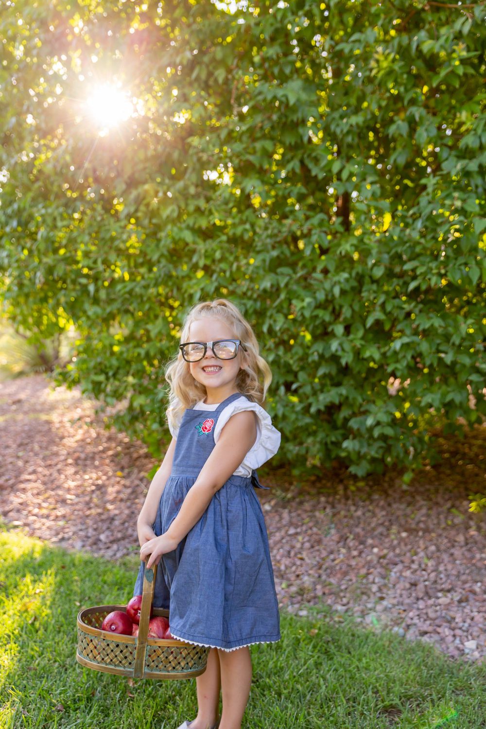 Child in denim dress holding basket outdoors with sunlight streaming through trees.
