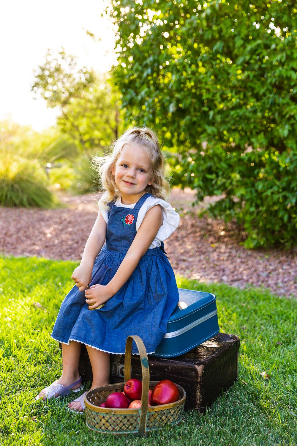 Student in blue dress sits on vintage suitcases with basket of apples outdoors.