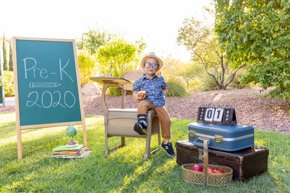 Student poses playfully on vintage desk next to Pre-K chalkboard and stacked suitcases.