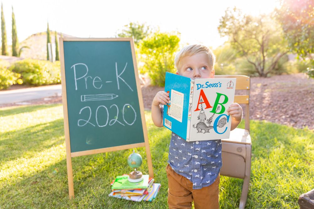 Student peeks from behind ABC book next to Pre-K chalkboard sign outdoors.