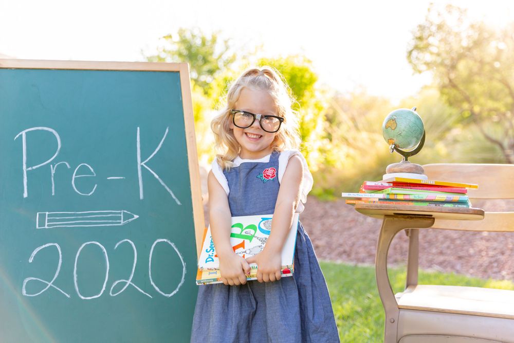 Student standing by chalkboard with books and globe for Pre-K 2020 photo in sunny garden.