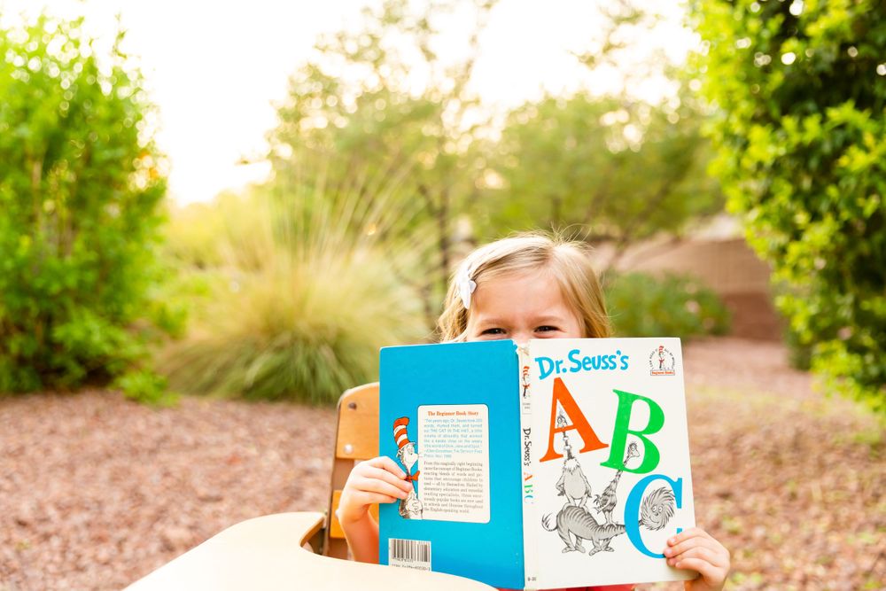 A child poses with an ABC book in front of green foliage on a sunny day.