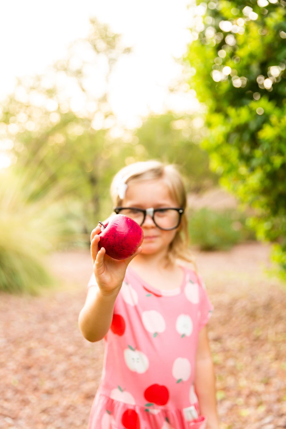 A student in a pink polka dot dress holds up a bright red apple outdoors in natural lighting.