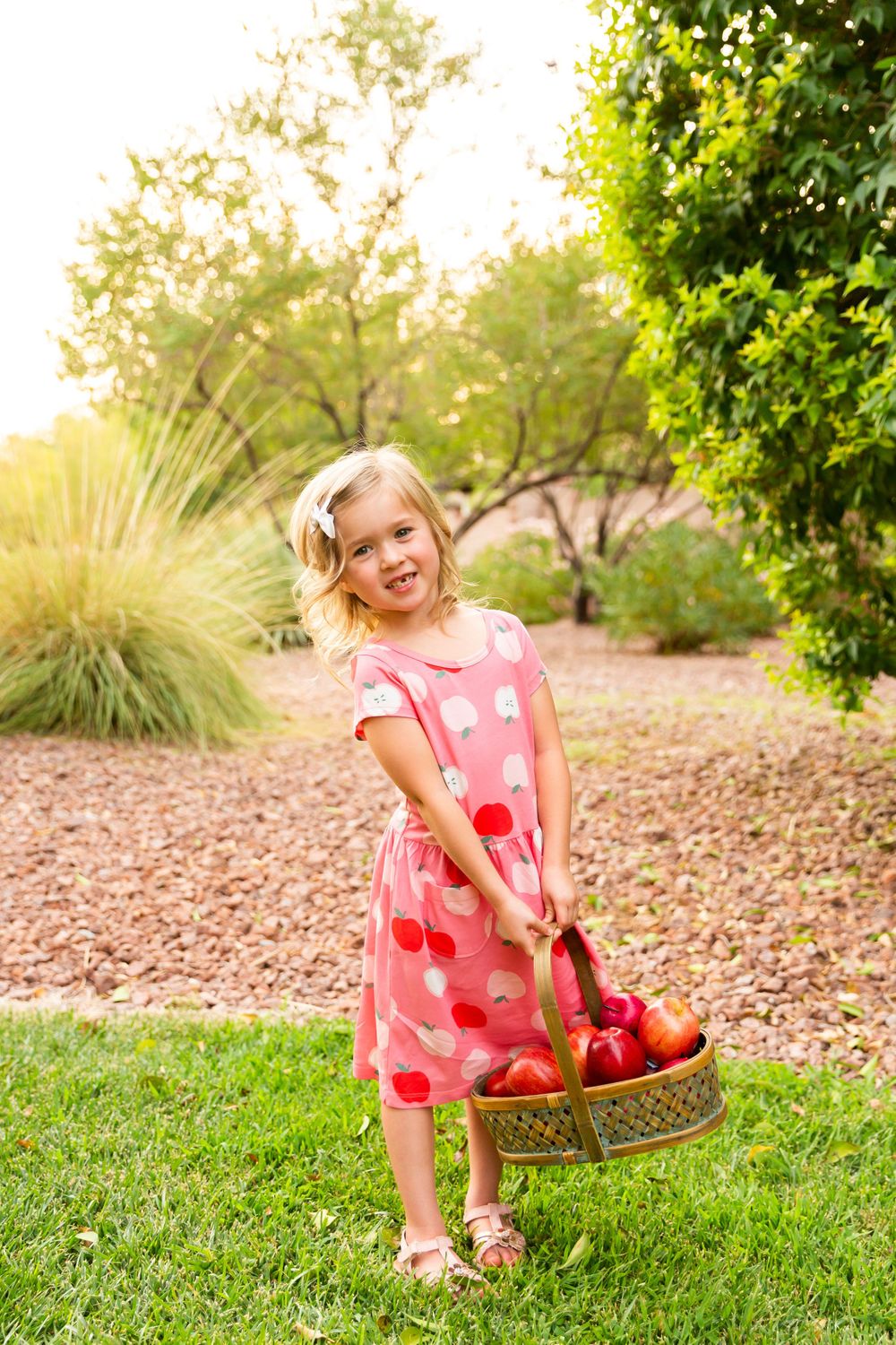 Child in pink polka dot dress holds basket of apples in sunny garden setting.