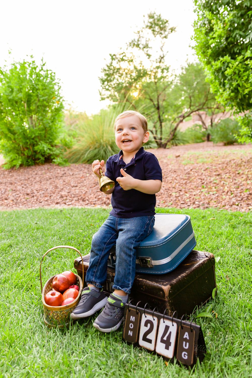 Happy child sits atop vintage suitcases next to countdown blocks and basket of apples in garden.