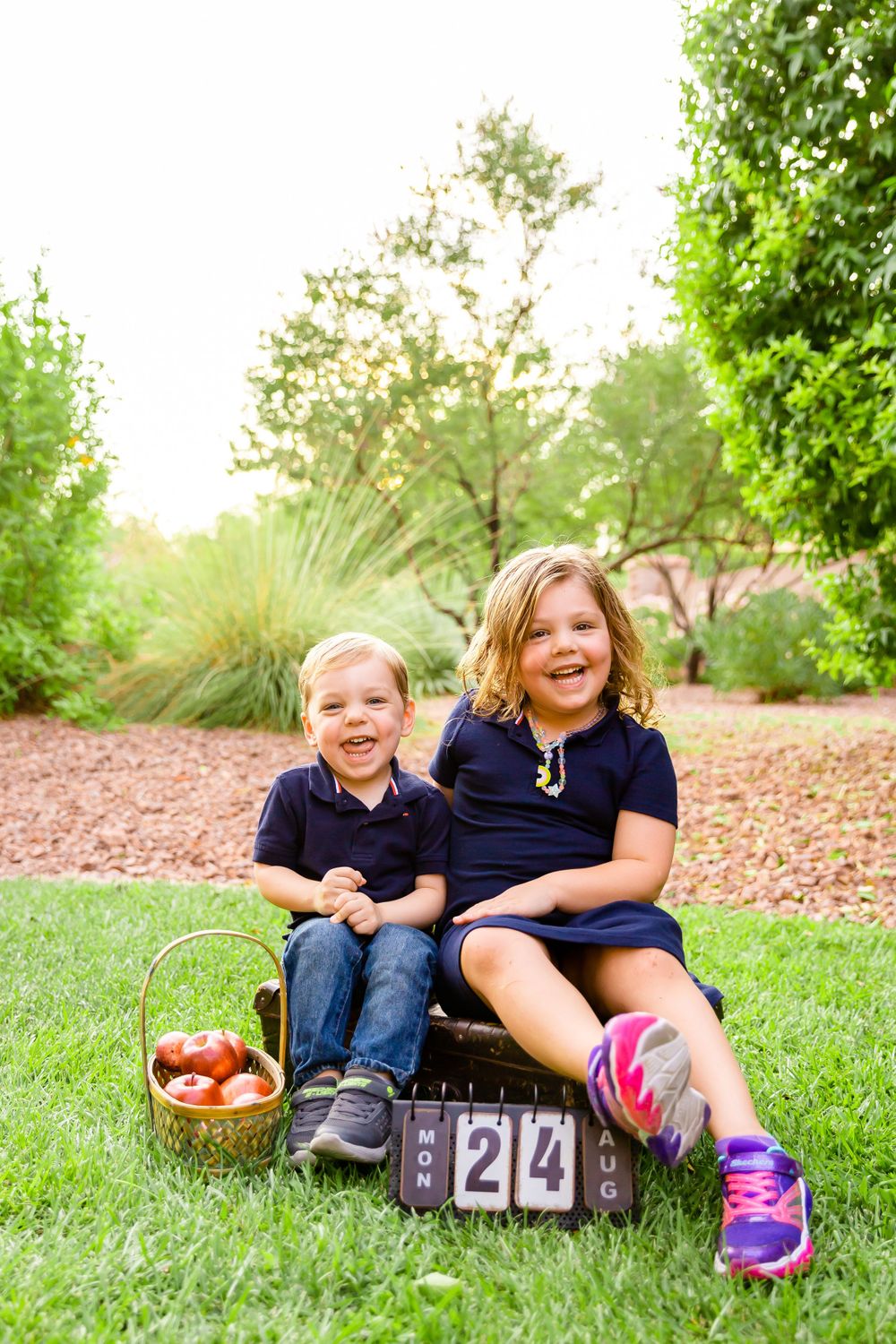 Two children pose on vintage luggage with apples and countdown blocks in a lush garden setting.