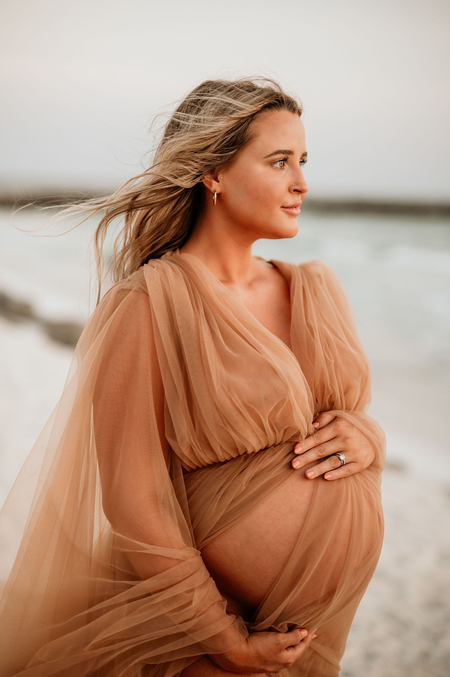 Maternity portrait in flowing peach dress on beach with wind-swept hair at sunset.