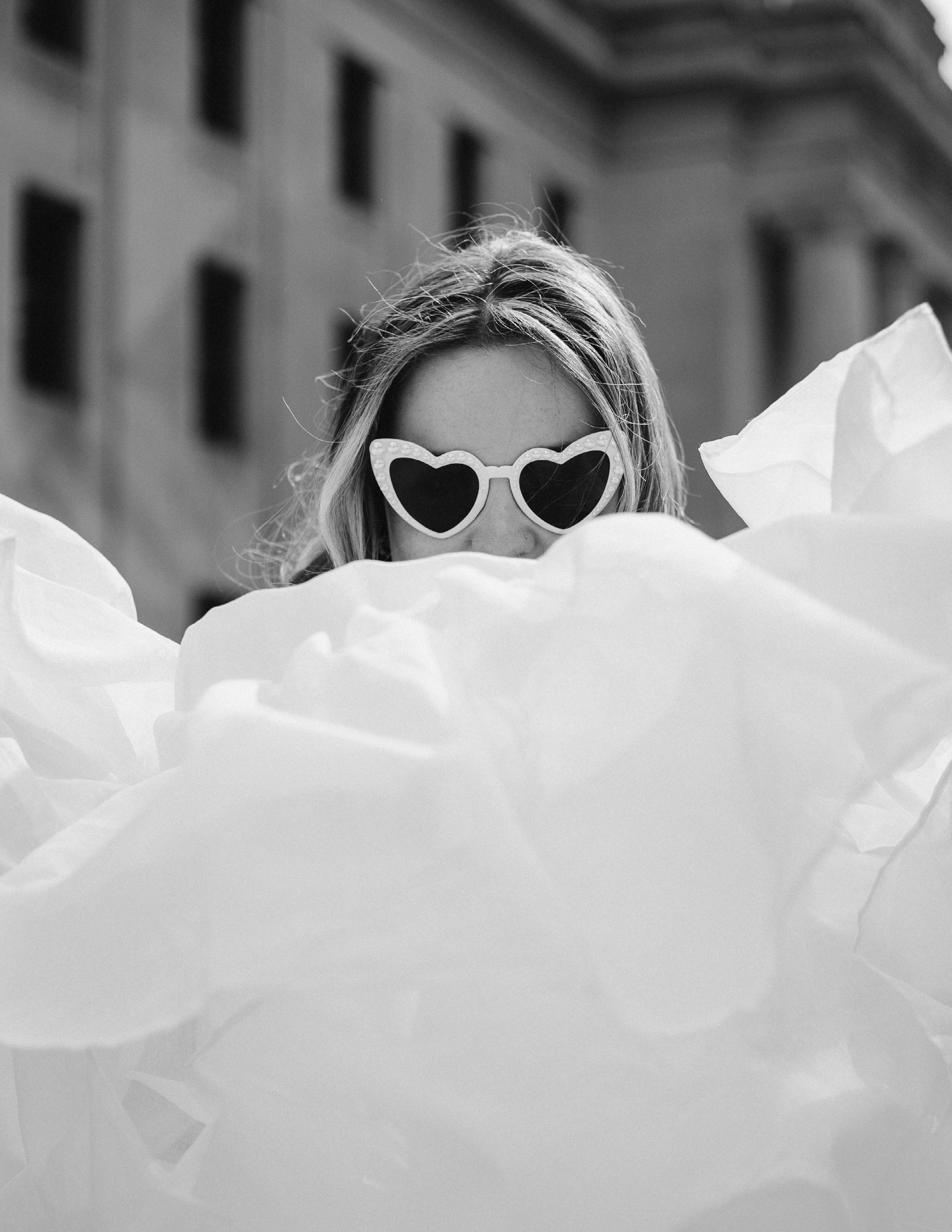 Black and white photo of someone wearing heart-shaped sunglasses peeking through white tulle fabric.