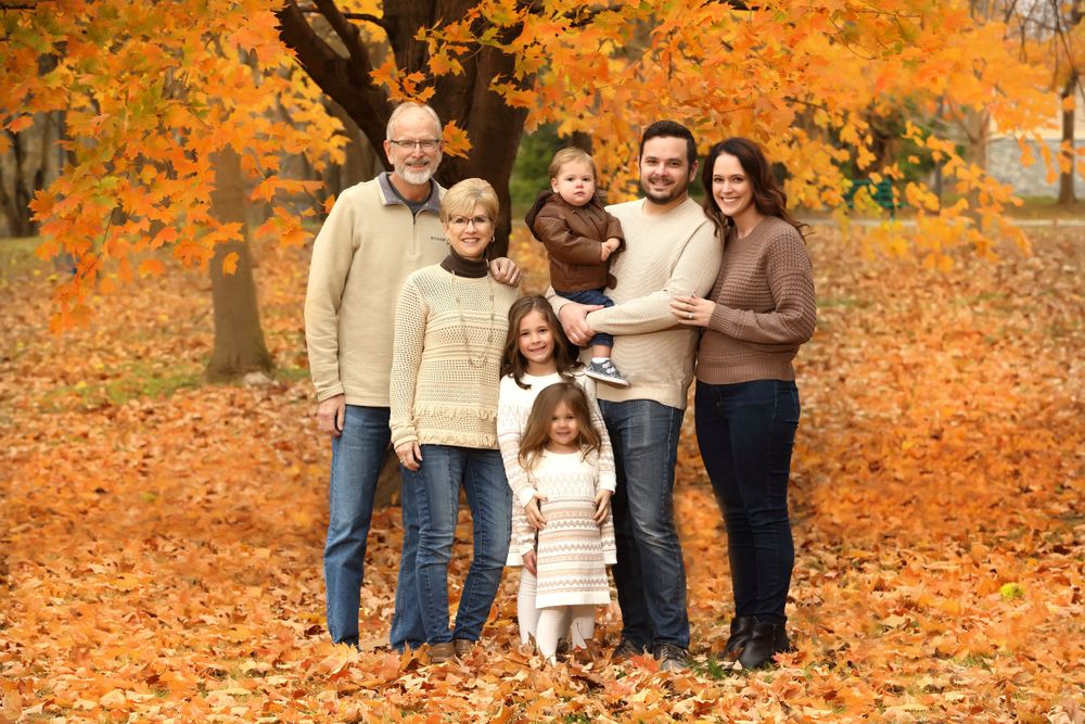 Multi-generational family poses together on fallen autumn leaves with vibrant orange trees in background.
