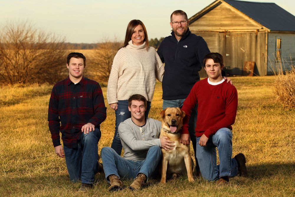 Family poses outdoors with their dog in front of rustic farmhouse at golden hour.