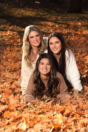 Three women in light colored sweaters pose together among fallen autumn leaves.