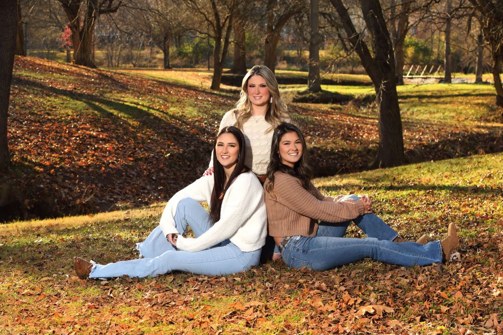 Three friends in casual winter clothing sitting together among fallen autumn leaves.